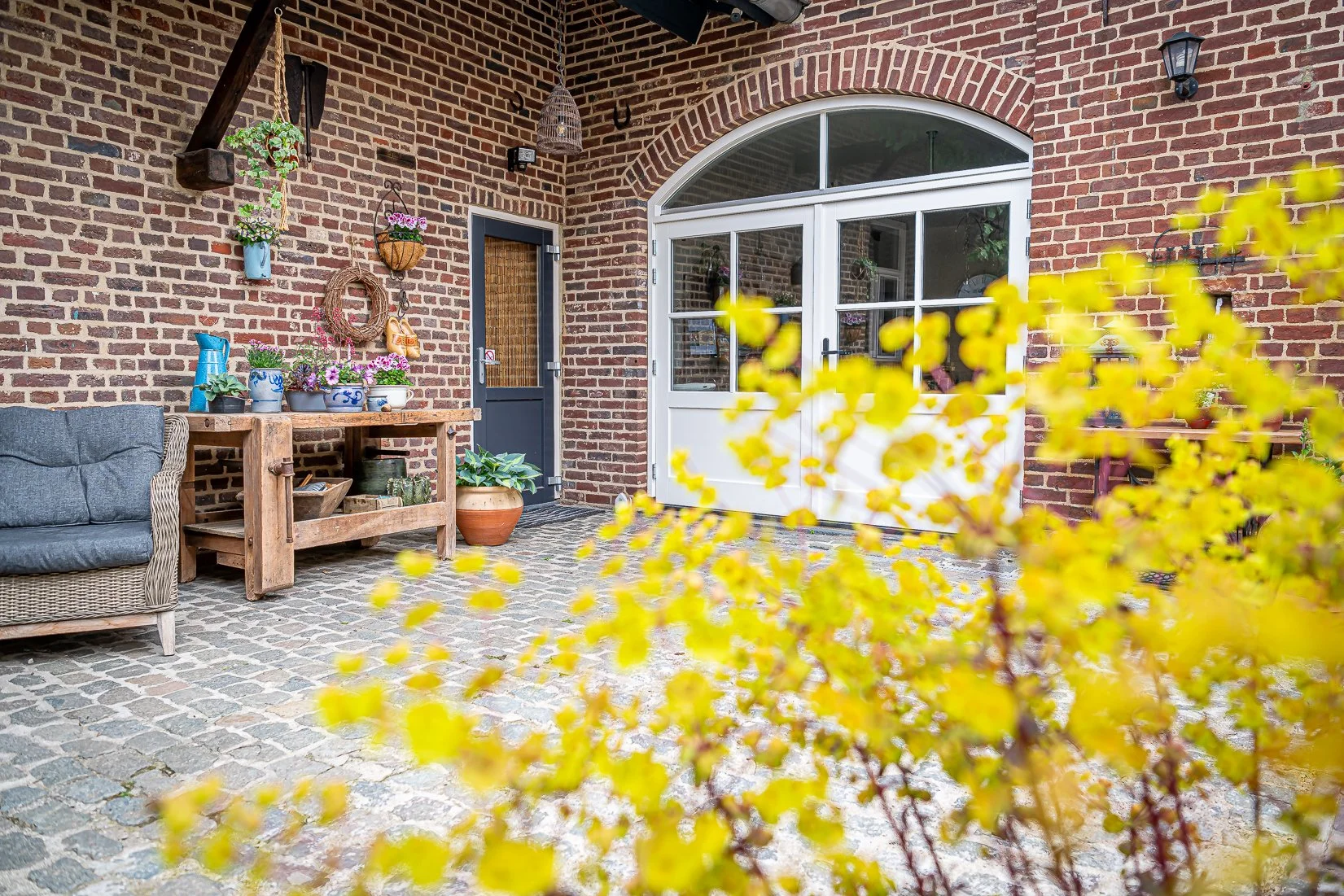 Een gezellig patio met een bakstenen muur, een bank met kussens, een houten tafel met planten en decoraties, en een witte deur met glas. In de voorgrond zijn gele bladeren van een struik zichtbaar.
