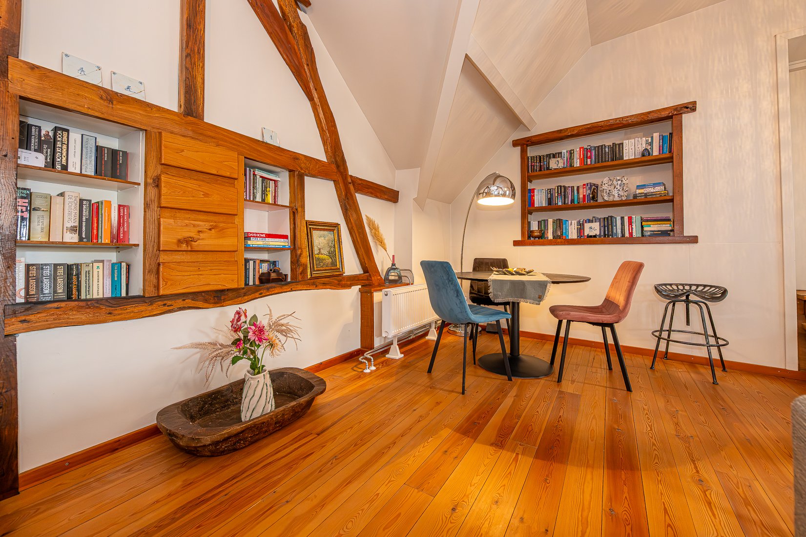Een gezellige woonkamer met houten vloeren en ingebouwde boekenplanken. Er staat een ronde tafel met drie stoelen, en er is een vloerlamp. Op de vloer ligt een grote houten botaniek met een vaas bloemen. De muren en het plafond hebben een lichte tint