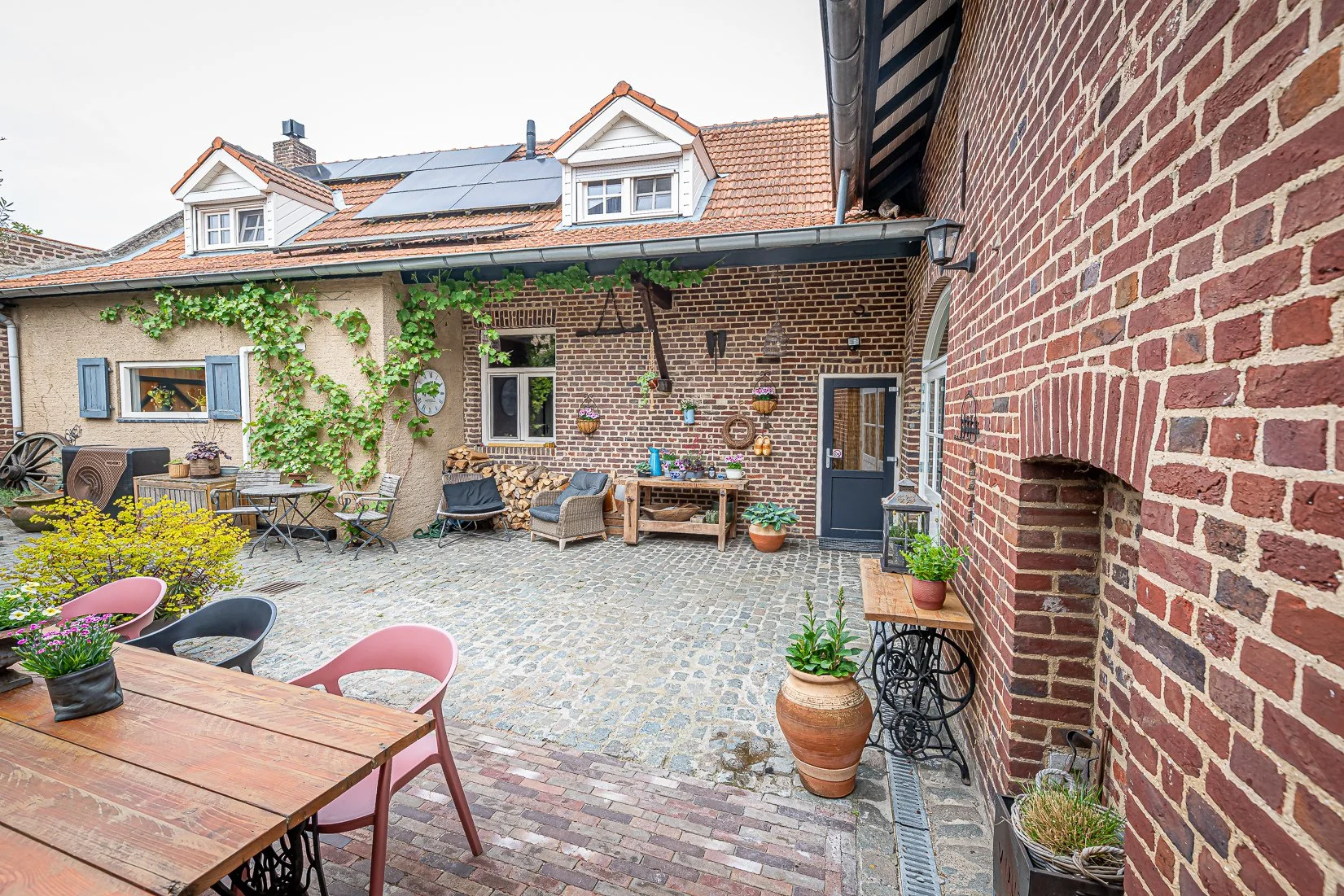 Een gezellige binnenplaats met tuinmeubilair, planten en decoraties, omgeven door bakstenen muren en een huis met zonnepanelen op het dak.