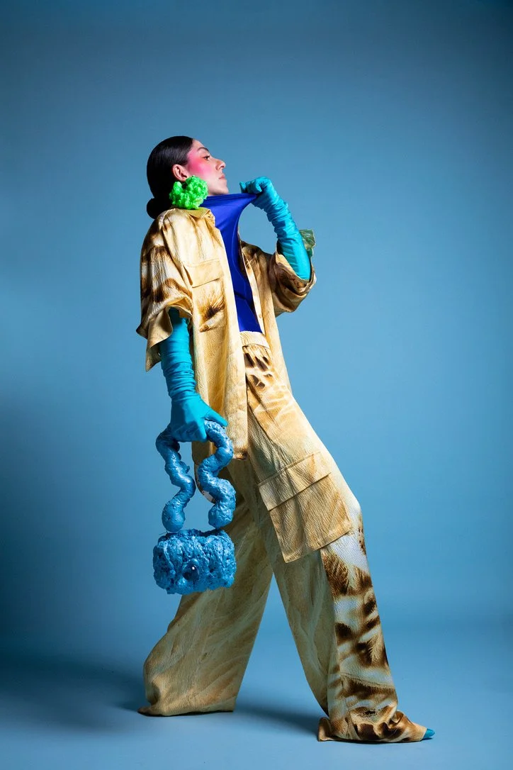 A woman wears a gold and brown patterned outfit with bright blue gloves, holding a blue textured object, against a plain blue background.