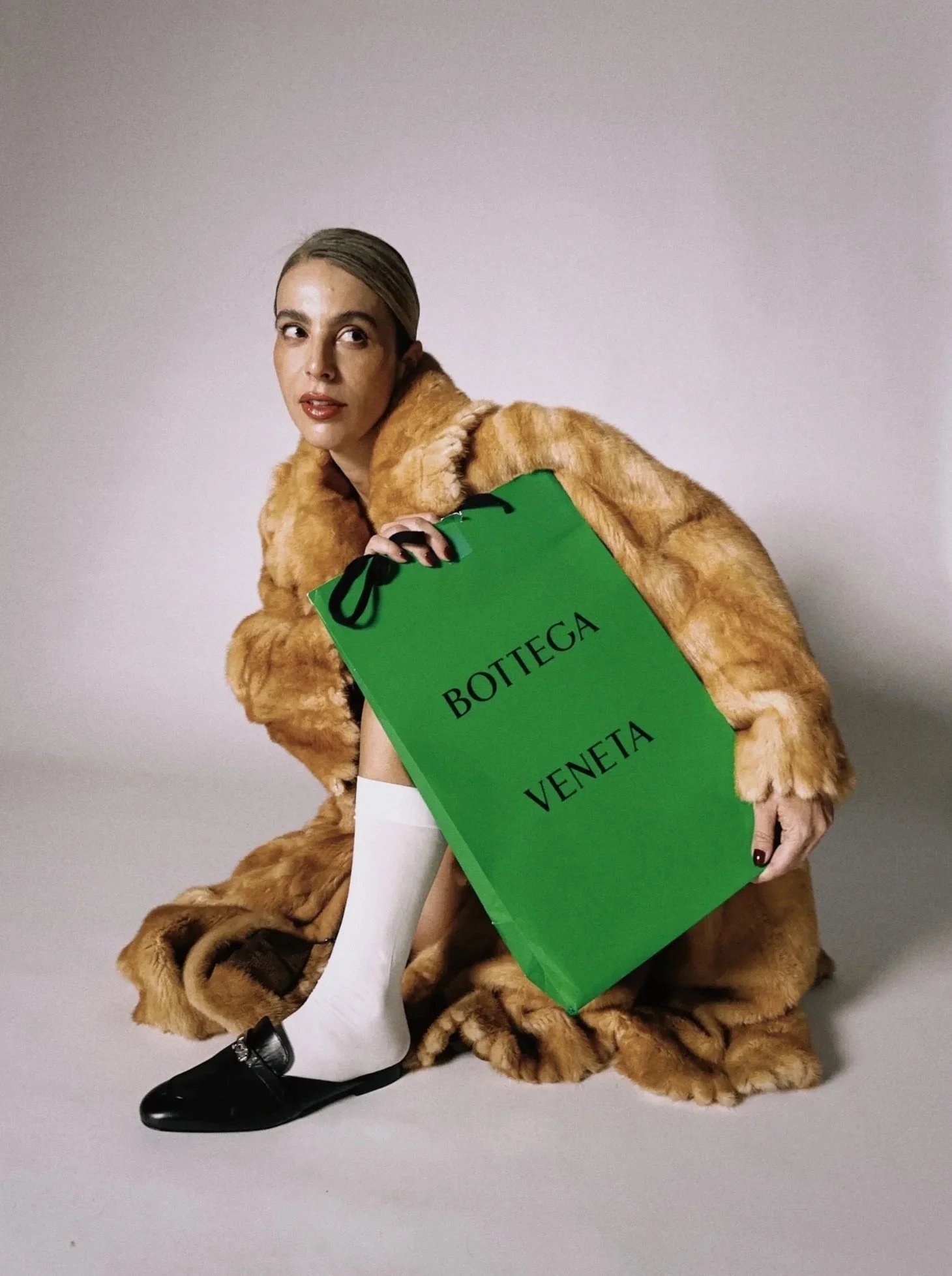 A person with long hair and makeup, wearing a faux fur coat, white thigh-high stockings, and black shoes, holding a green shopping bag with "BOTTEGA VENETA" written on it, sitting against a plain background.