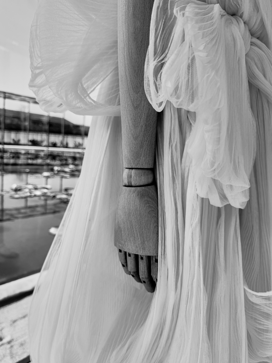 Close-up of a mannequin's wooden hand and part of a flowing, pleated dress with layered ruffles, with a parking lot and building visible outside the window.