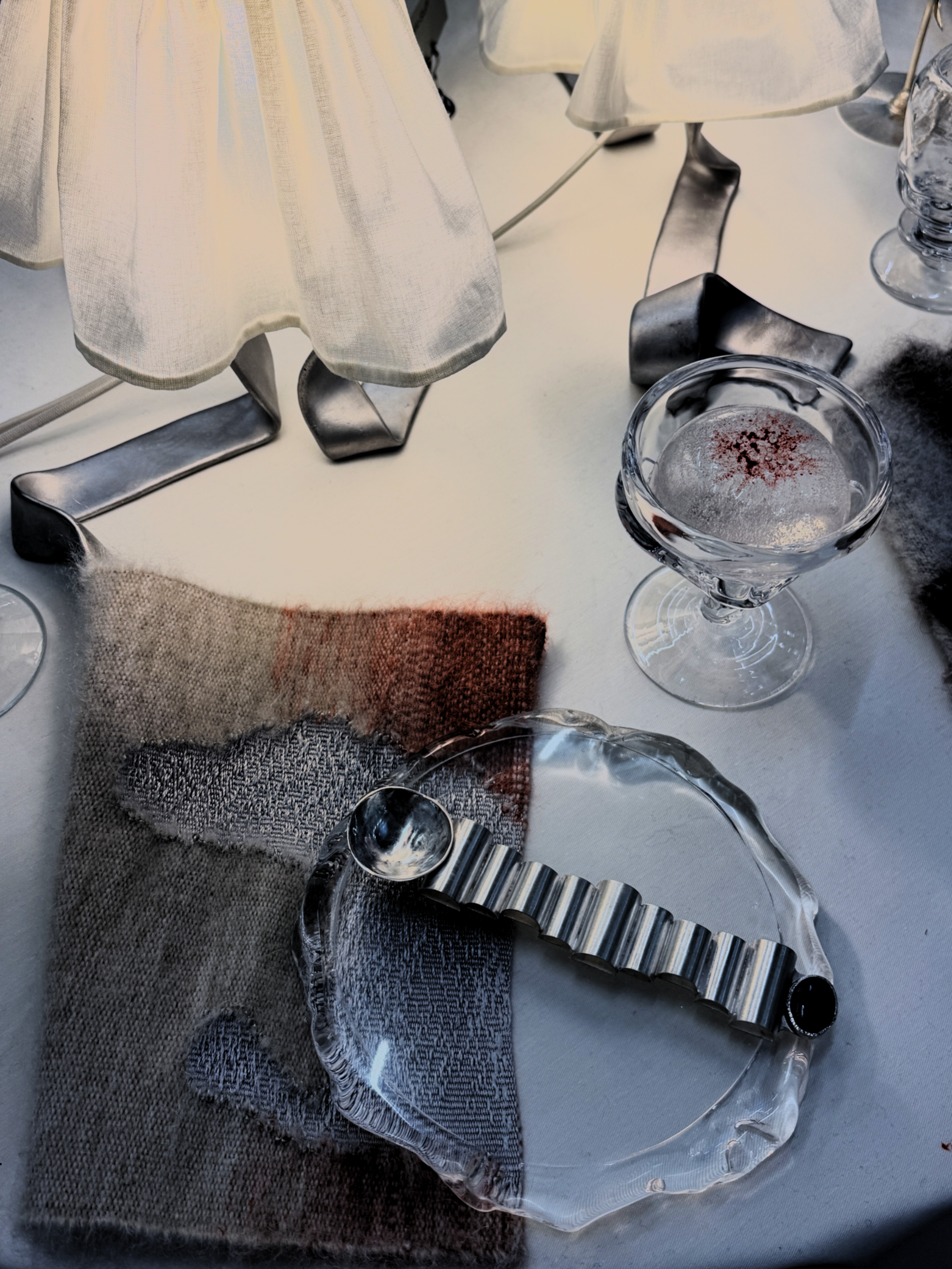 Close-up of a table setting featuring a clear glass dessert bowl with red sprinkles, metallic and glass serving utensils, textured cloth napkins, and a cream-colored lampshade, with a white tablecloth.