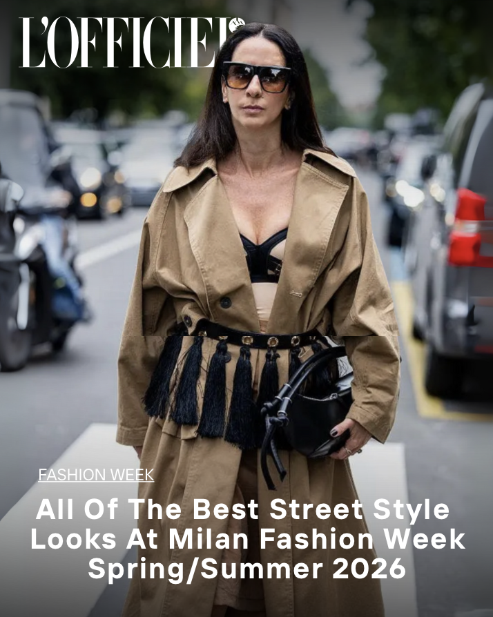 A woman with long dark hair wearing large sunglasses, a khaki trench coat draped over her shoulders, a black bra, and black tassel belt standing outdoors near parked cars, with a magazine cover headline about Milan Fashion Week Spring/Summer 2026.