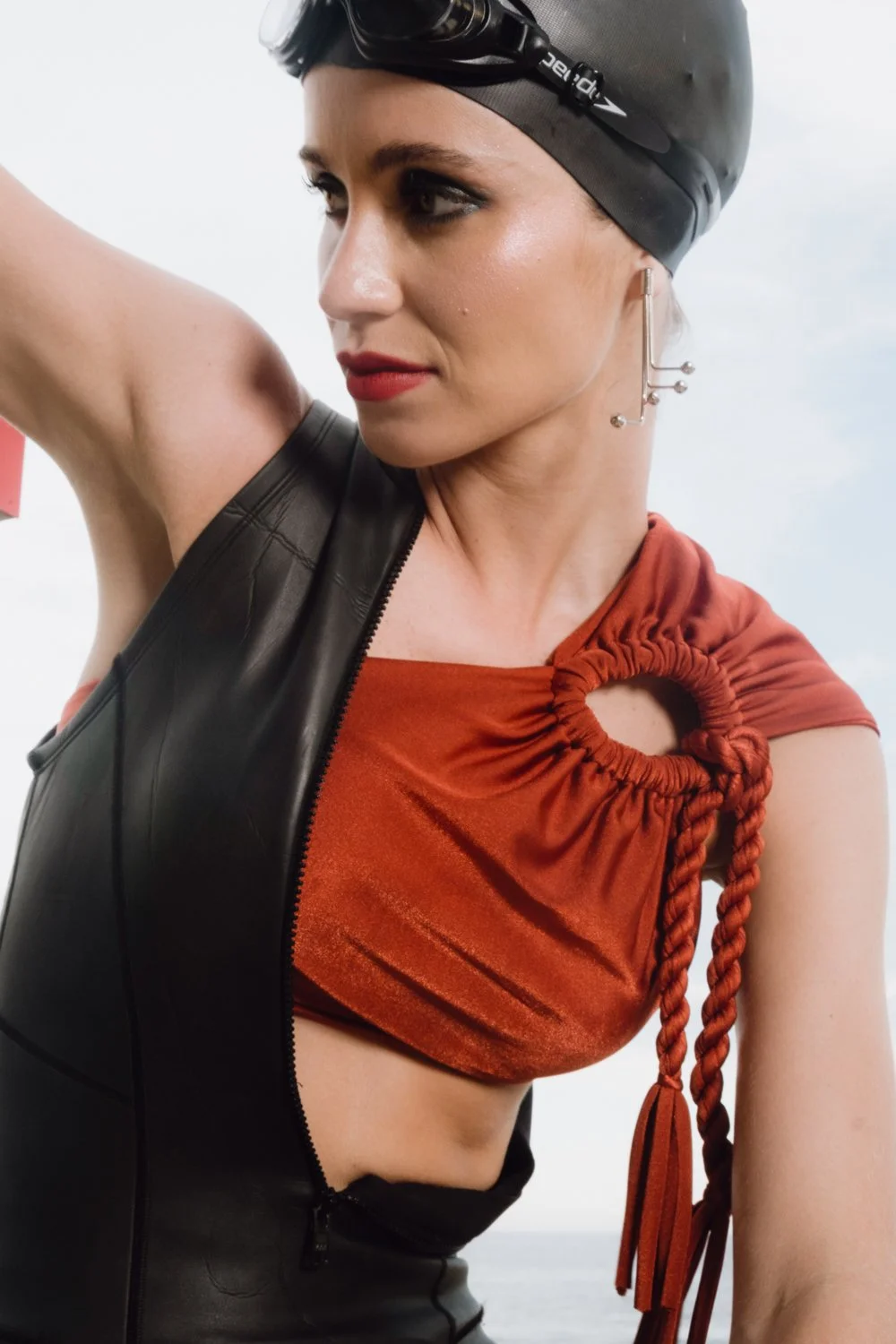 A woman wearing a black swimming cap, black goggles on her forehead, and a sleeveless black leather vest over an orange top with a large circular cutout and braided details. She has makeup with red lipstick and is looking to her left, outdoors with a