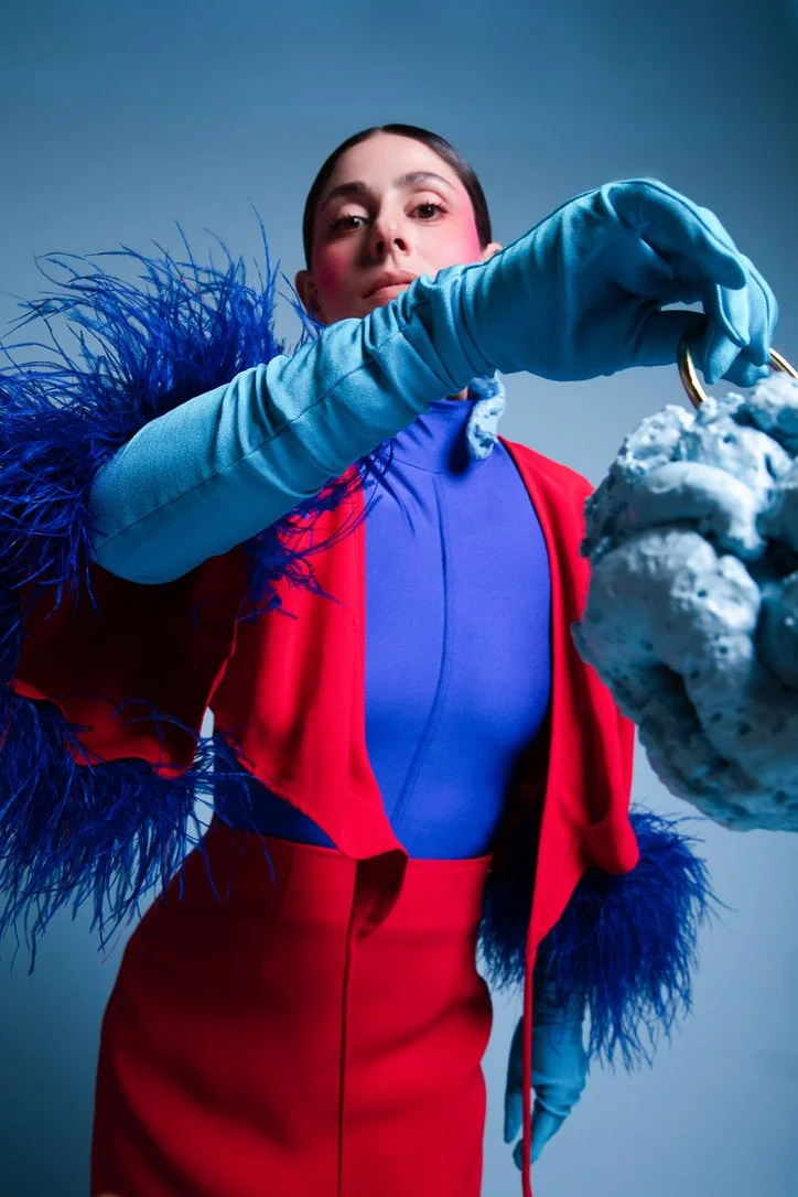 A woman wearing a colorful outfit with a blue top, red skirt, red jacket, and long blue gloves poses while holding a large textured object with a hook.
