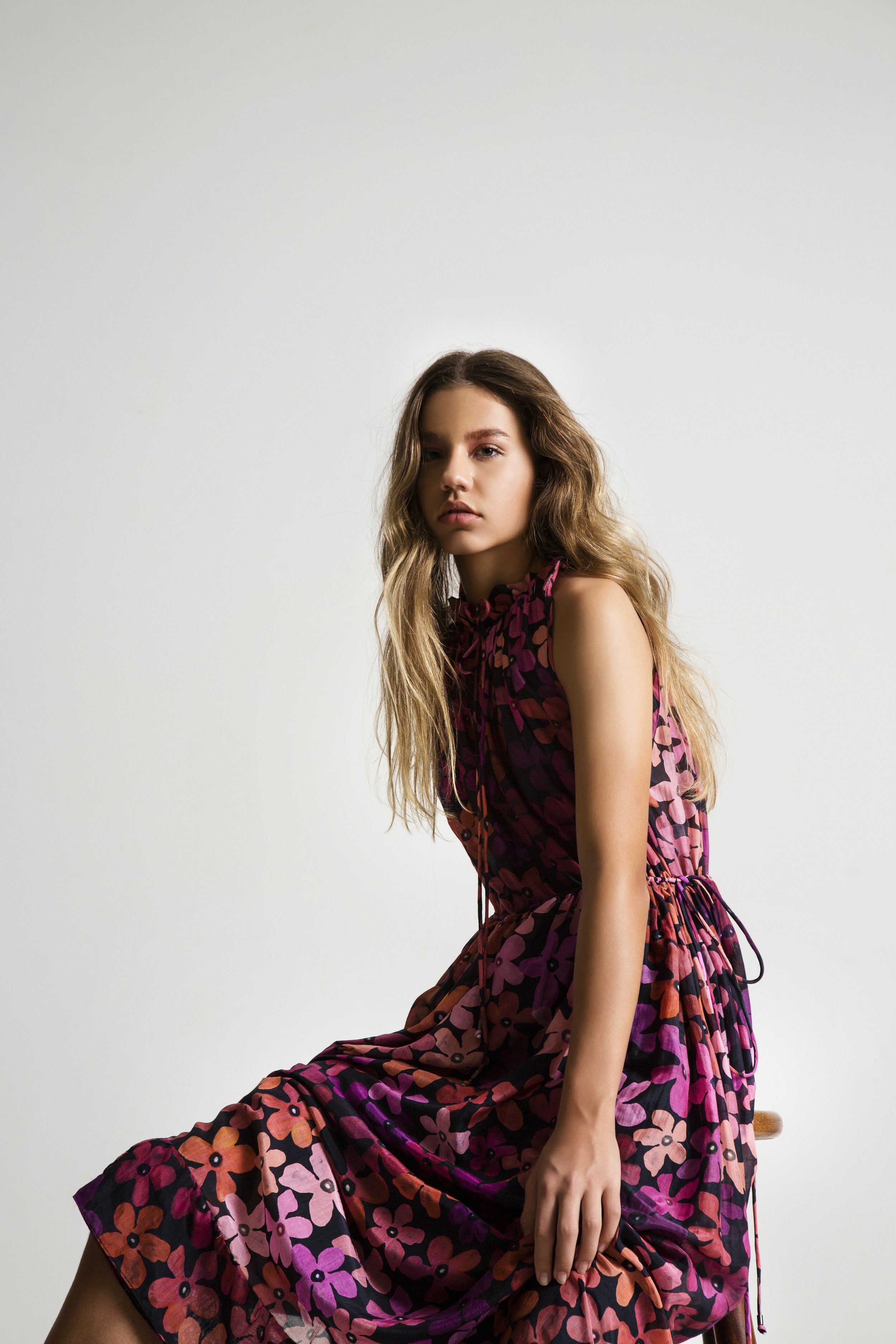 A young woman with long wavy hair sitting on a wooden stool against a plain white wall. She's wearing a sleeveless, patterned dress with shades of pink, orange, purple, and black, featuring a floral or abstract design. The woman has a neutral express