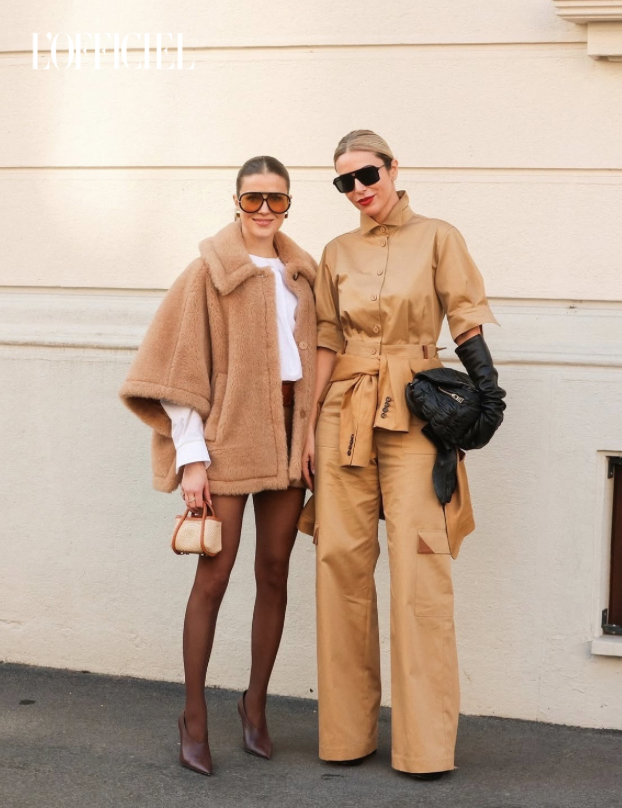 Two women standing outdoors against a beige wall, wearing fashionable outfits; one has on a tan fuzzy coat, shorts, sheer tights, and high heels, and the other is in a tan jumpsuit with black gloves and sunglasses.
