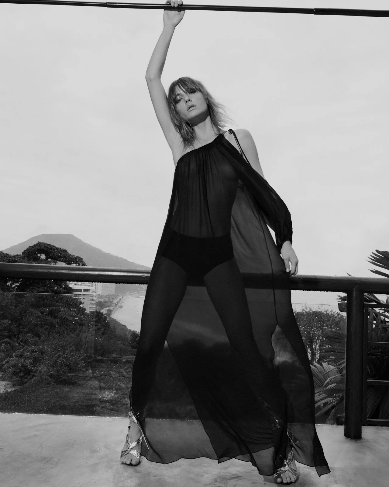 A woman stands on a balcony holding a pole above her head with one hand. She is wearing sheer black clothing and high heels. The background shows a landscape with mountains and trees, and the image is in black and white.