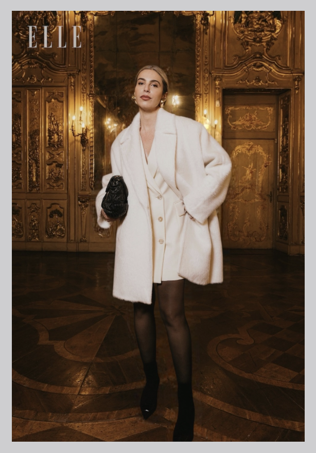 A woman, Flavia Millen,  dressed in a white MAX MARA coat holding a black DELVAUX clutch purse, standing in an ornate gold-adorned room with intricate woodwork and lighting fixtures.