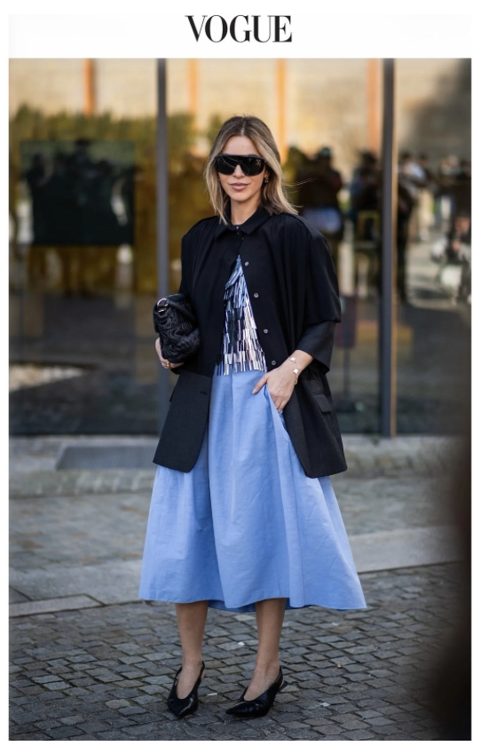 Woman standing outdoors in front of a glass building, wearing sunglasses, a black blazer over a patterned blouse, a long light blue skirt, and black heels, holding a black clutch. The Vogue logo appears at the top.
