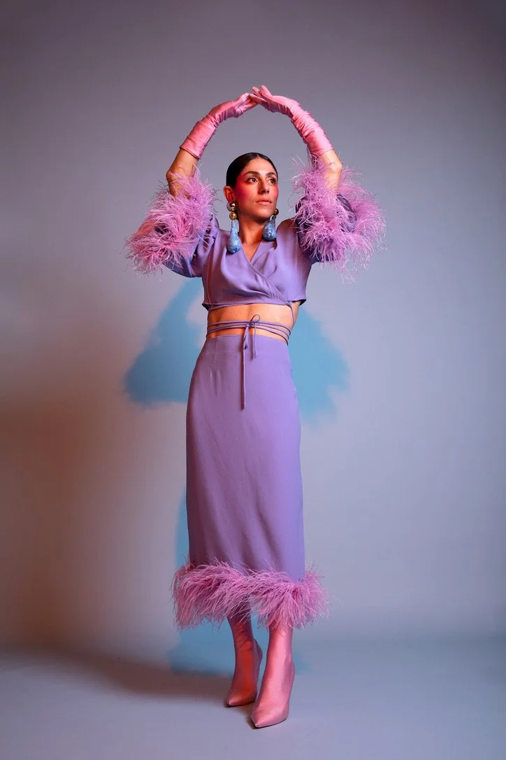 A woman dressed in a purple two-piece outfit with feathered pink trim on the sleeves and hem, wearing pink gloves, earrings, and pink high heels, standing against a gray background.