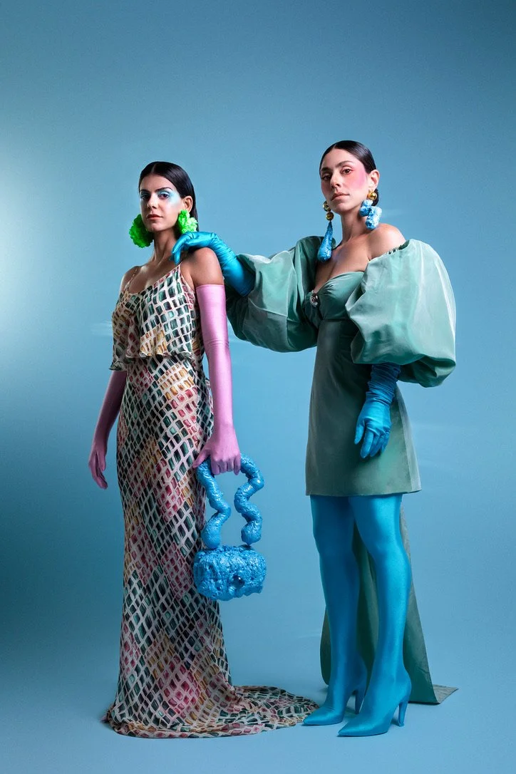 Two women posing against a plain background, wearing colorful, avant-garde fashion with bold accessories and gloves, showcasing a high-fashion editorial look.