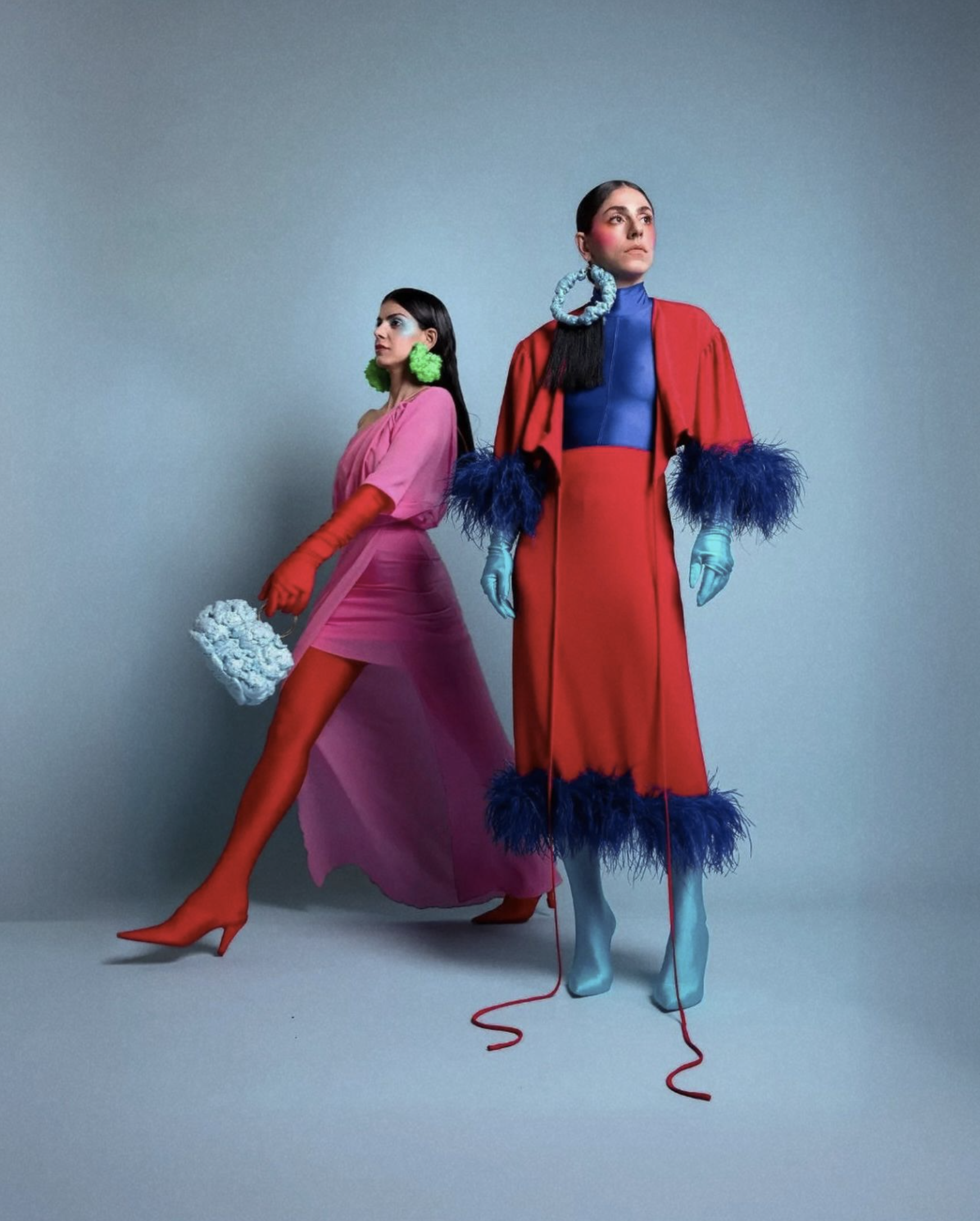 Two women dressed in colorful, avant-garde outfits with bold makeup and large earrings standing against a plain background.