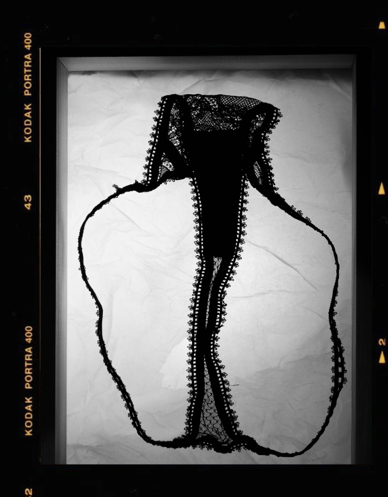 A black and white sketch of a nude woman from the back, with her hair up, on crumpled paper, framed by a vintage camera viewfinder.