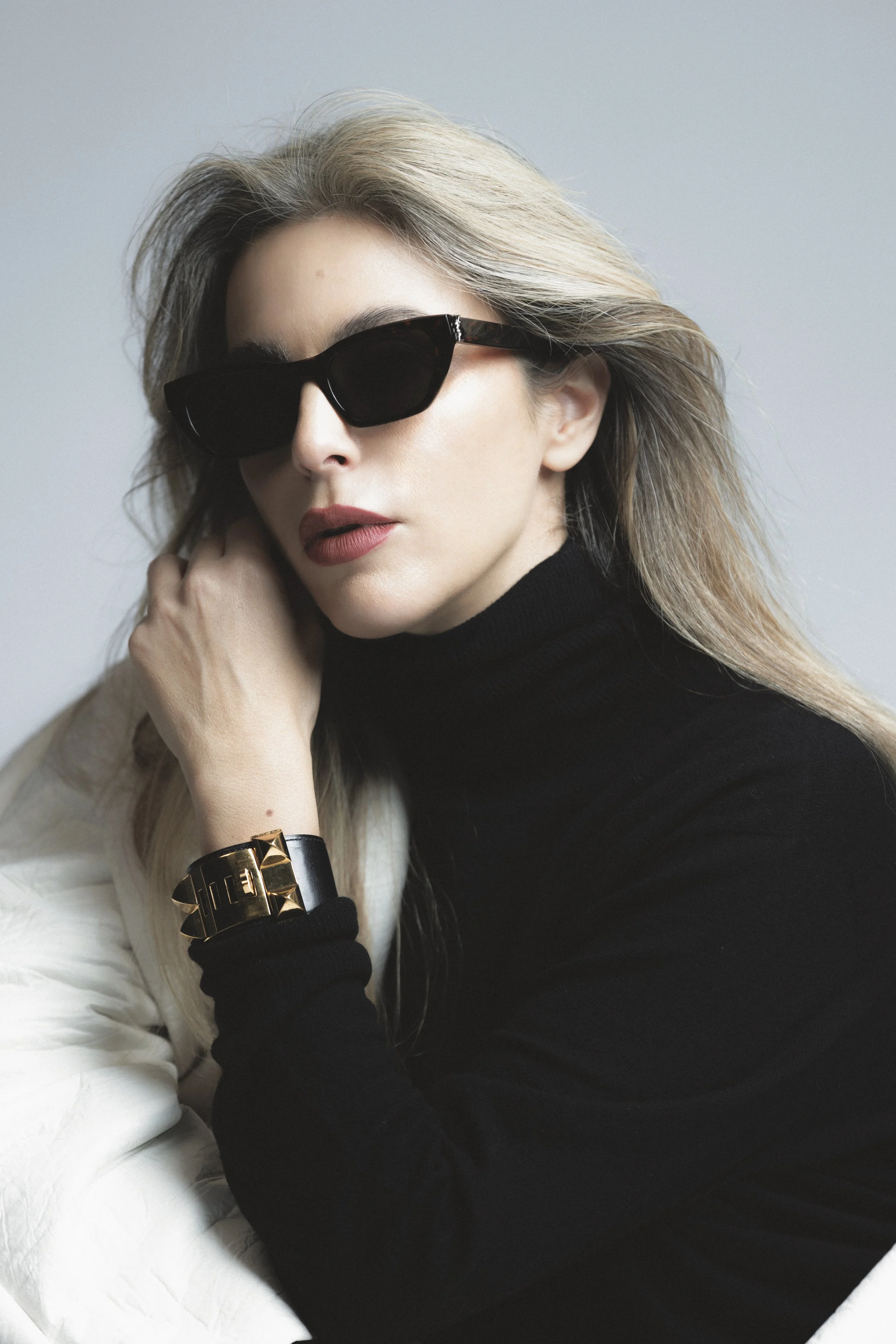 A woman with blonde hair wearing black sunglasses, a black long-sleeve top, a black bracelet, and a gold watch, sitting on a white quilted surface, with her hand near her face.