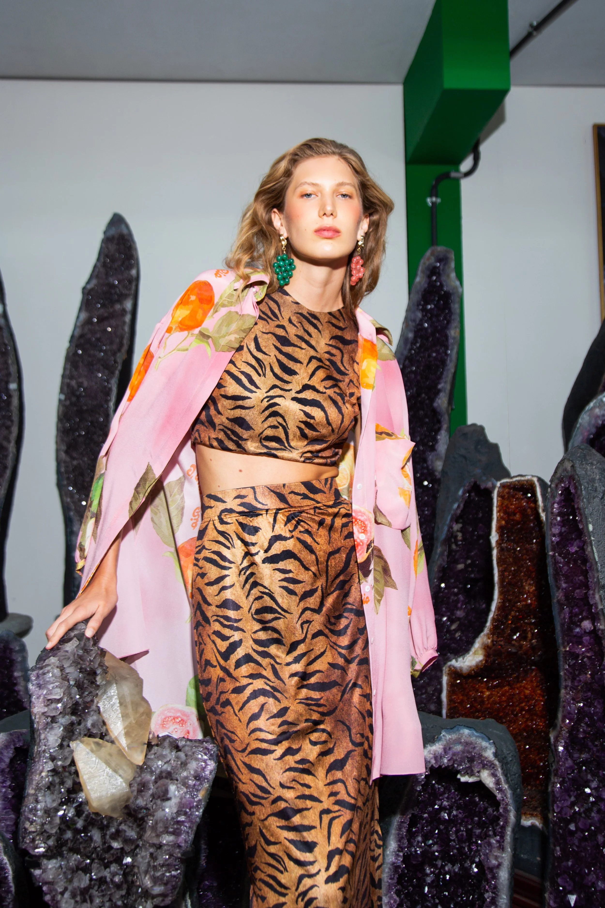 A woman in animal print clothing with large green and pink earrings stands in front of amethyst and other mineral geodes.
