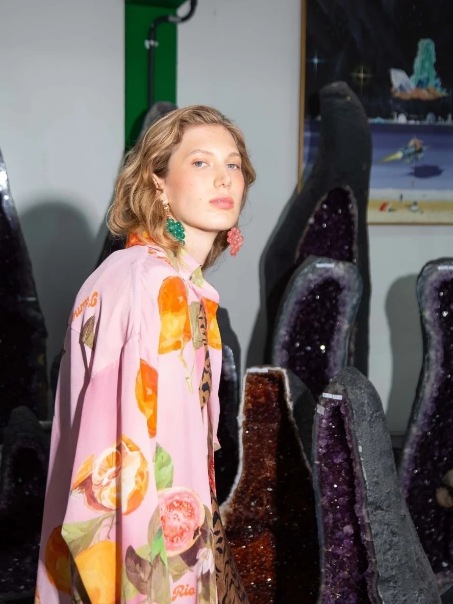 A woman with wavy, shoulder-length blonde hair and fair skin posing among large, purple, geode-like crystals. She is wearing a pink patterned shirt with orange, green, and pink flower designs; she has green and pink grape-shaped earrings. A colorful 