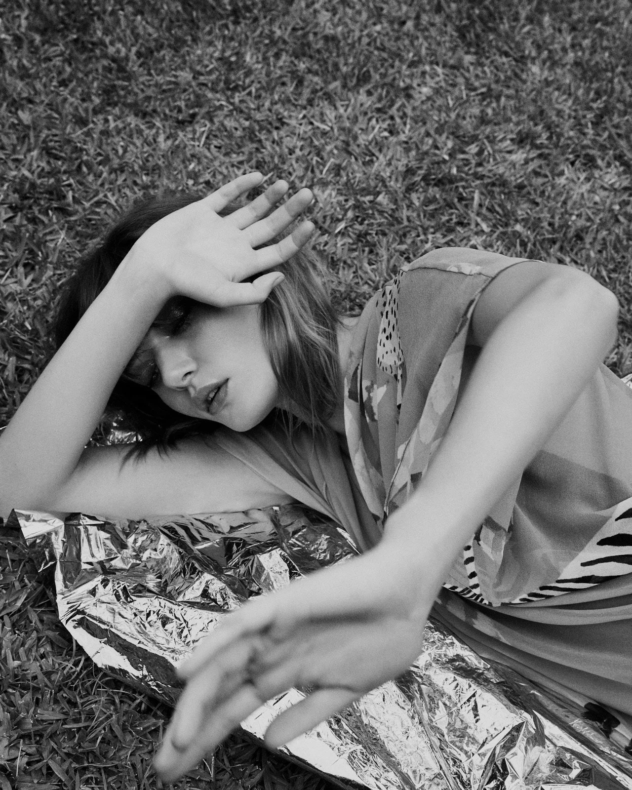A woman lying on grass, covering her face with one hand, with a reflective foil blanket under her. She has shoulder-length hair and is wearing a patterned top.