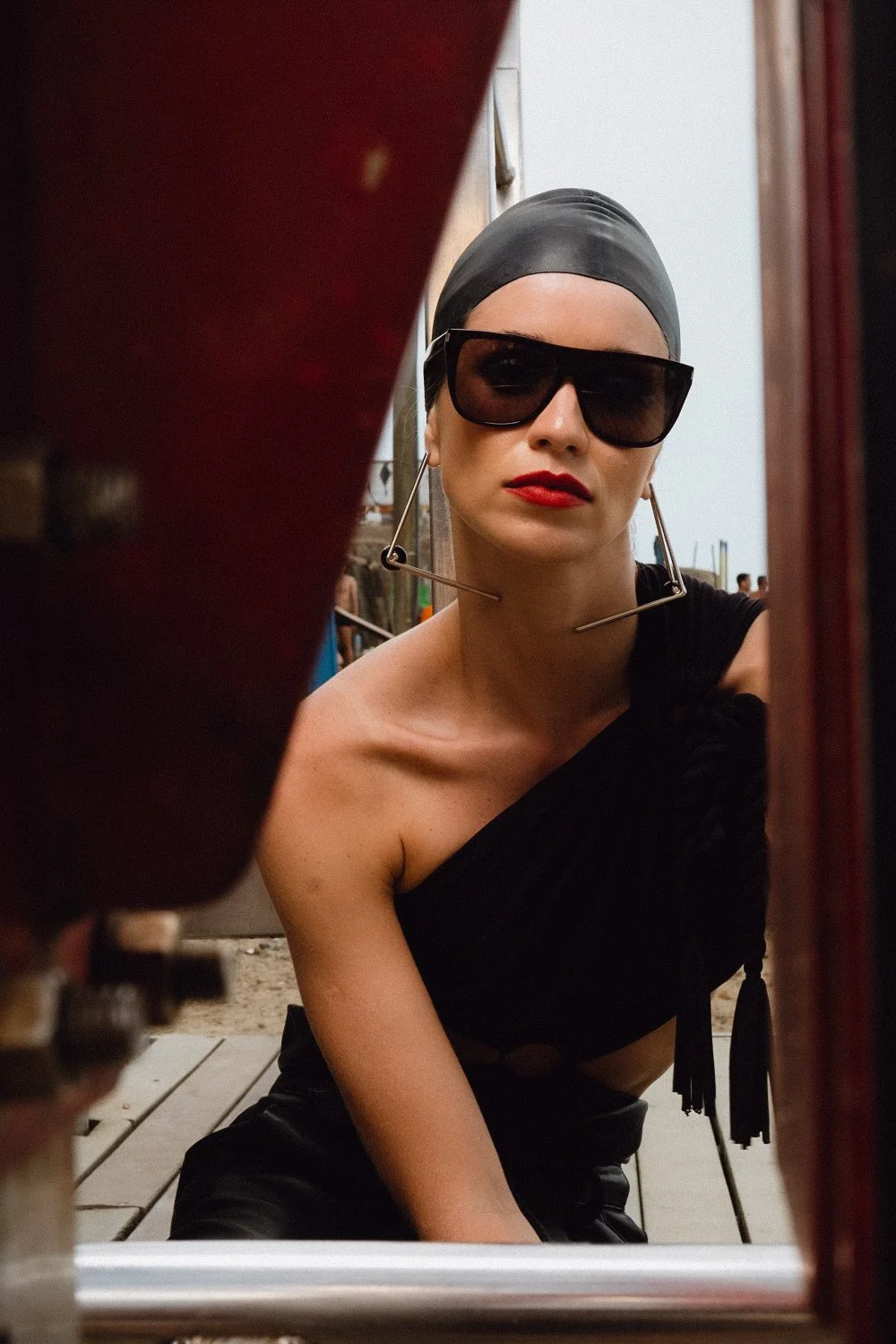 A woman with red lipstick, large black sunglasses, and a black headscarf is looking into a mirror at an outdoor setting. She is dressed in black, with one shoulder exposed.