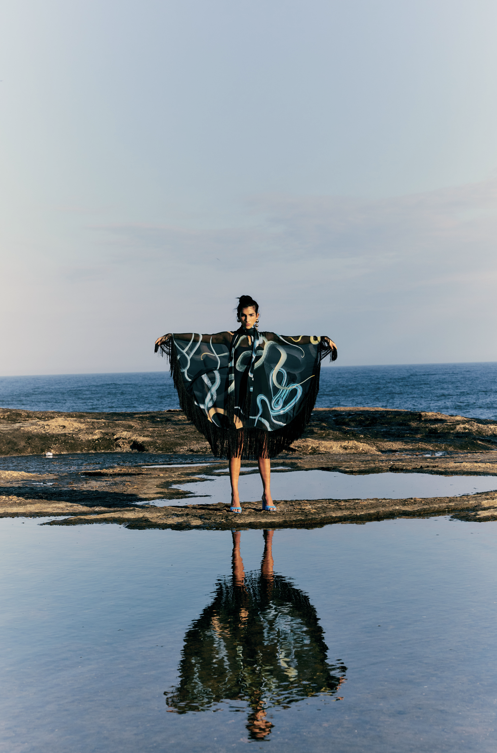 A woman wearing a black and colorful abstract patterned kimono and high heels stands on rocks by the ocean, with her arms outstretched holding the kimono open. Her reflection is visible in a pool of water on the rocks, with a cloudy sky above.
