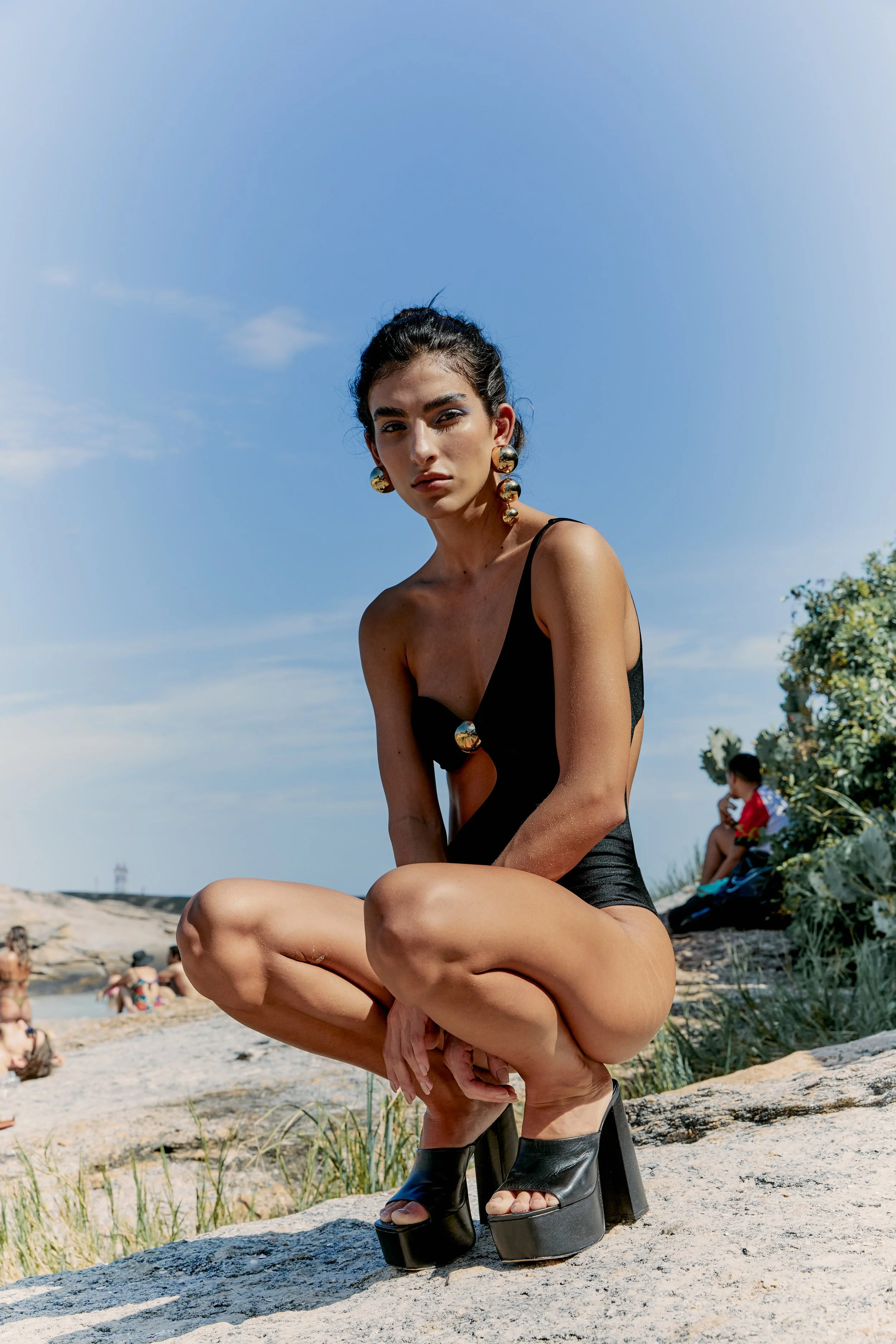 A woman with dark hair, wearing large gold earrings and a black swimsuit, is crouching on a rock at the beach against a blue sky. She is wearing black platform high heels and looking into the camera.