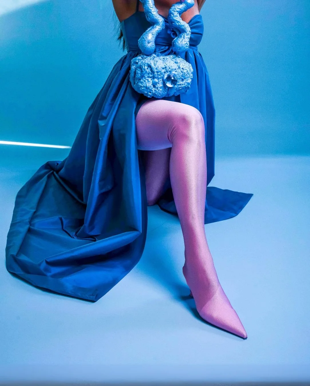 Person kneeling in a blue dress with a large blue textured handbag, pink high-heeled stocking, against a blue background.
