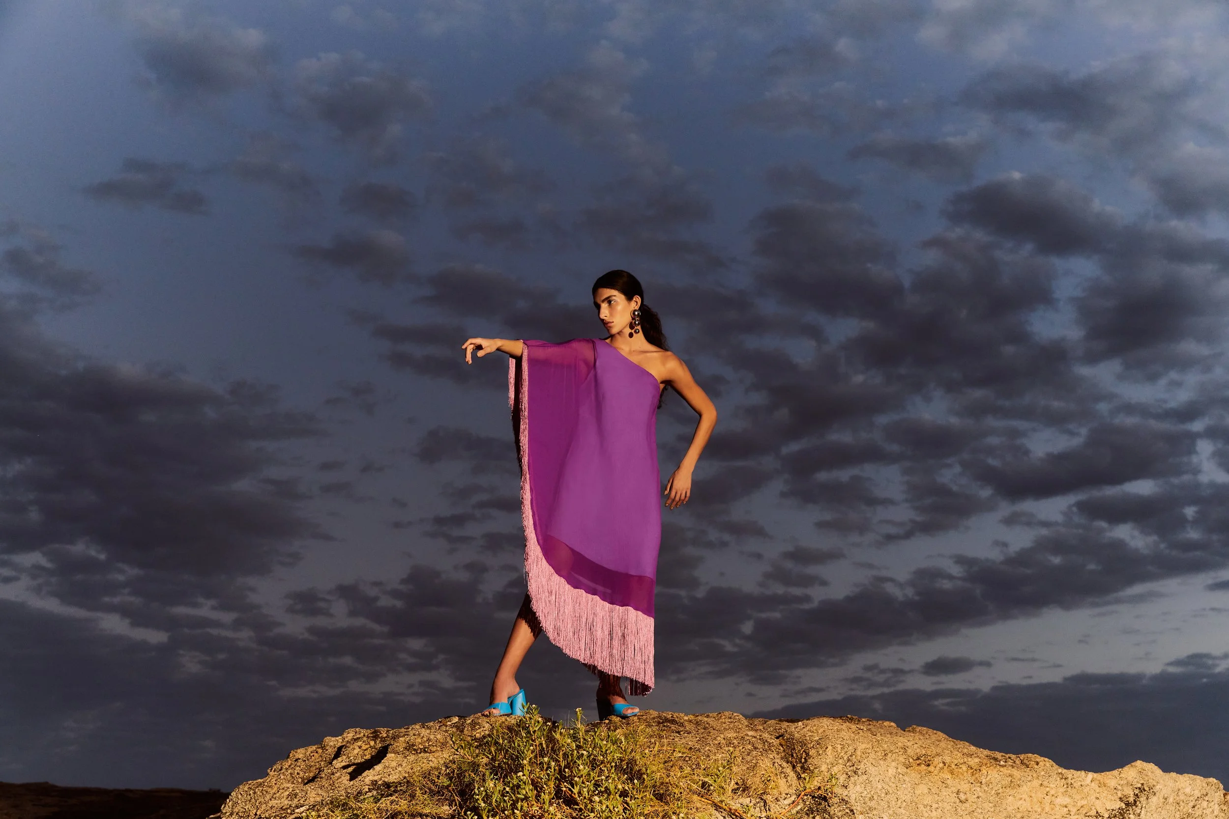 A woman in a purple dress standing on a rock with dark clouds in the background.