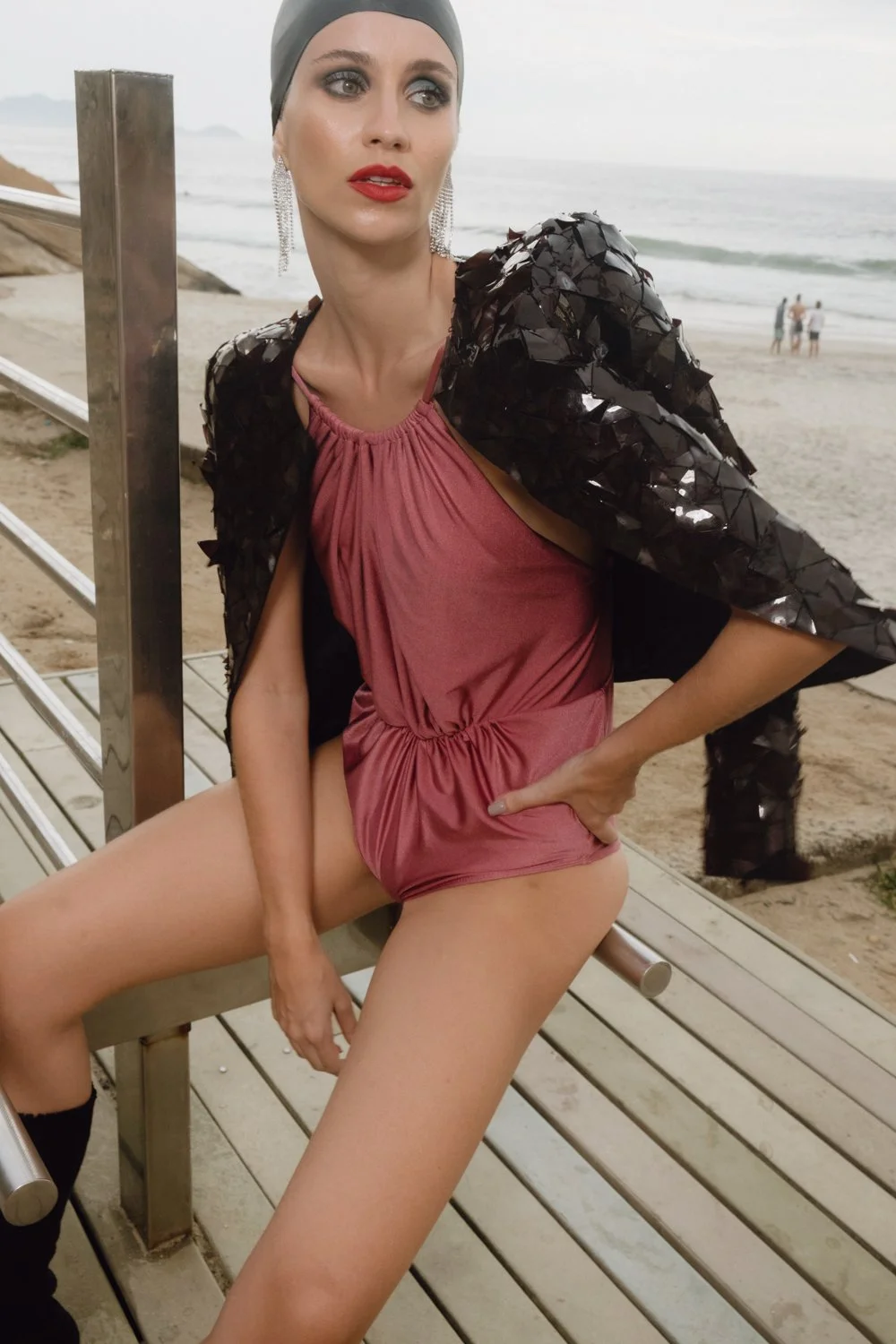 Woman with dramatic makeup and earrings posing on a beach promenade, wearing a pink satin dress and a black jacket with shiny black leaves on it.