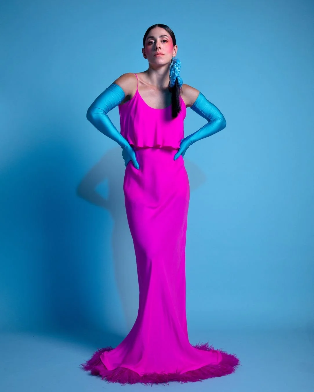 A woman wearing a bright pink evening dress with a looser top over a long skirt, paired with long blue gloves and a blue hair accessory, standing against a light blue background with her hands on her hips.