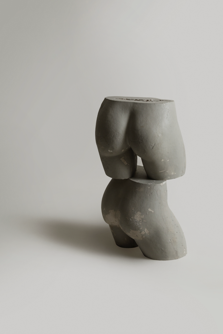 A modern abstract sculpture resembling a stylized human buttocks, made of gray material, on a light background.