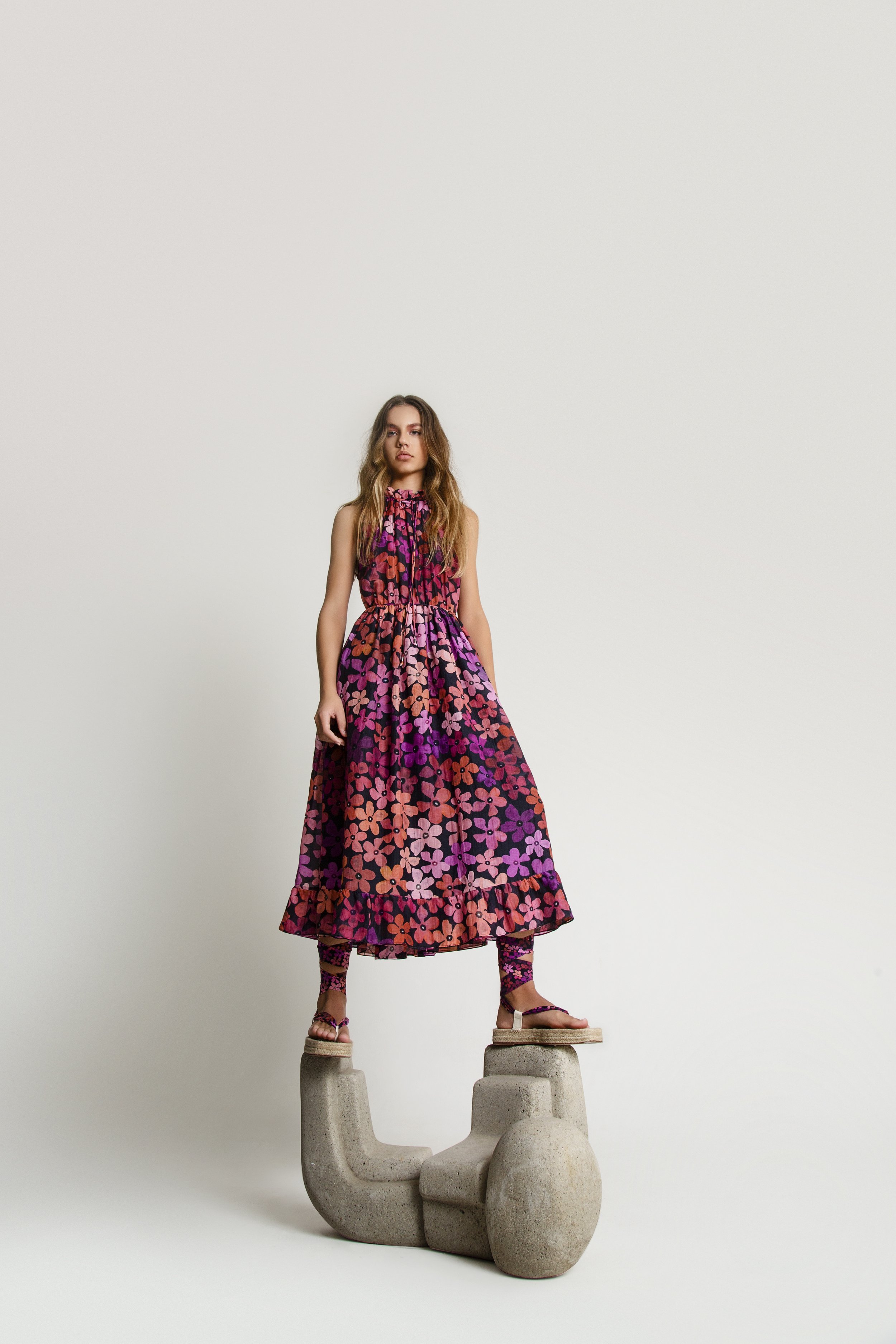 A young woman with long, wavy brown hair standing on multiple large concrete sculptures arranged in a creative pose against a plain white wall. She is wearing a colorful, floral-patterned maxi dress and matching sandals.