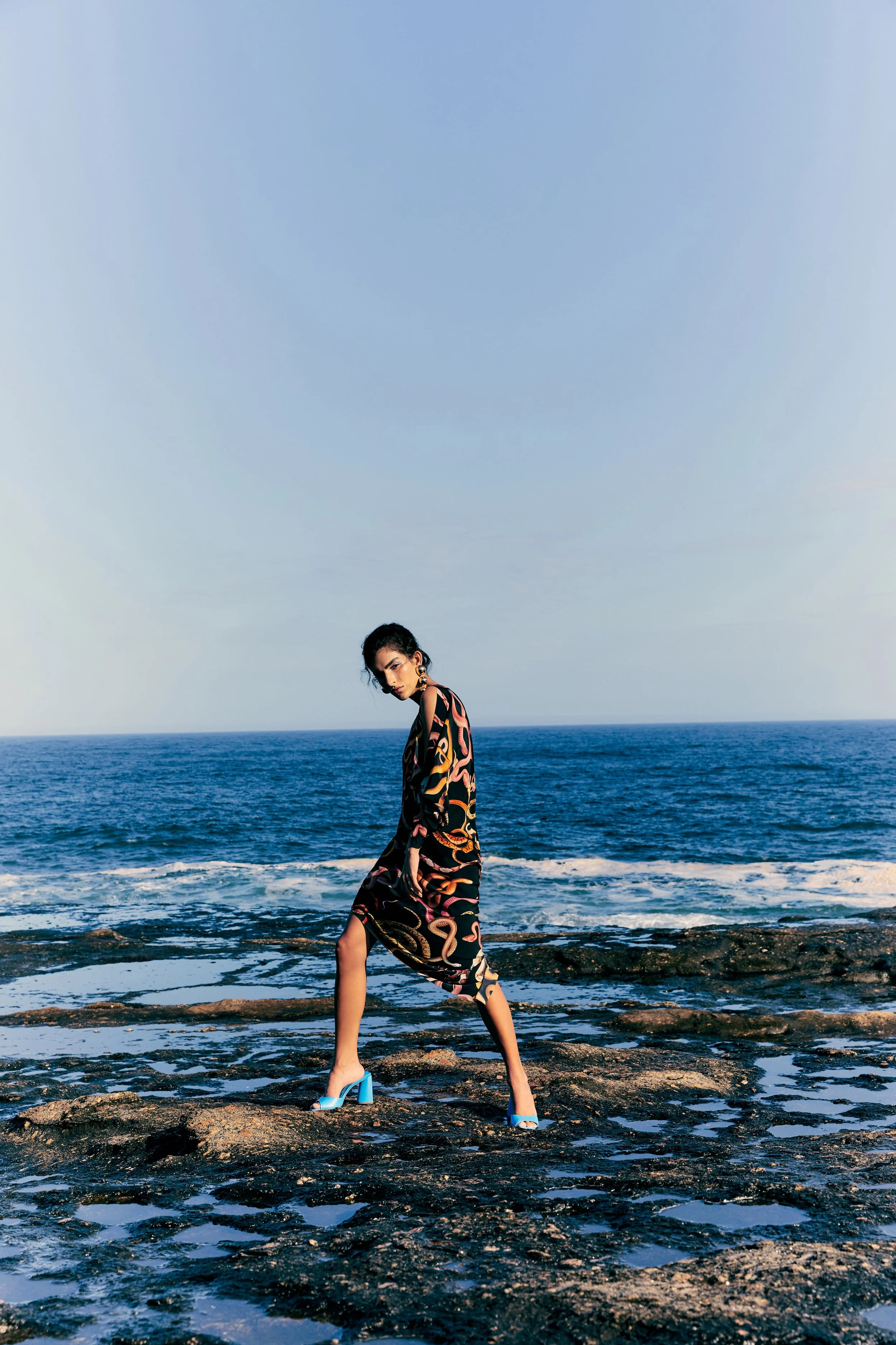 A woman standing on rocks by the ocean, wearing a colorful patterned dress and blue high heels, with the sea and sky in the background.