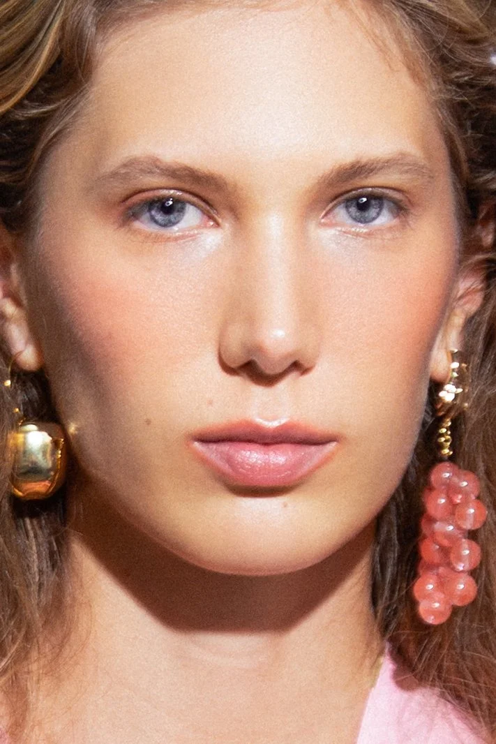 Close-up of a woman with blue eyes, light makeup, and gold earrings with pink beads.