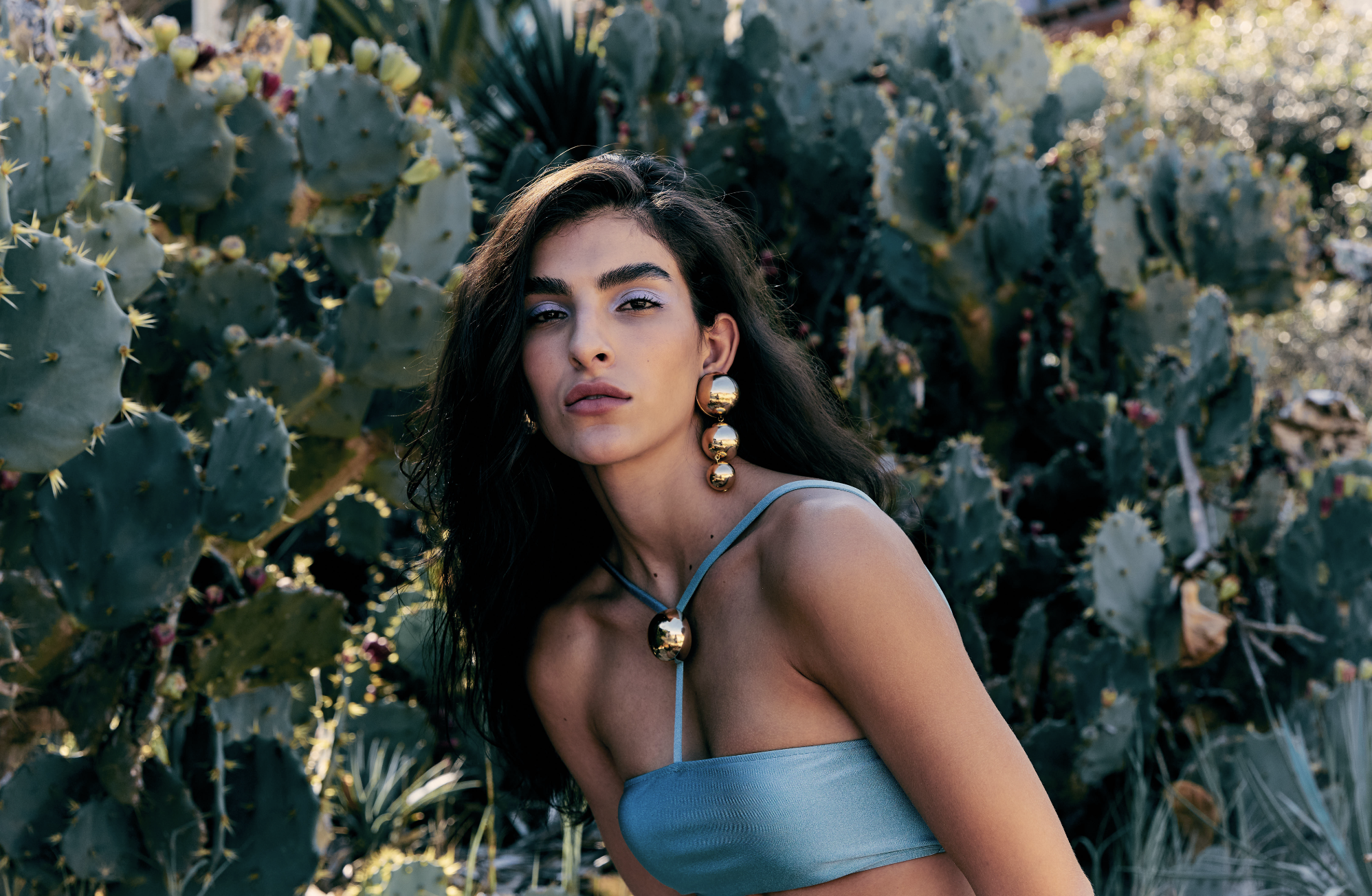 A woman with long dark hair poses in front of a cactus garden. She is wearing a light blue dress and large gold statement earrings and necklace. Sunshine filters through the background vegetation.