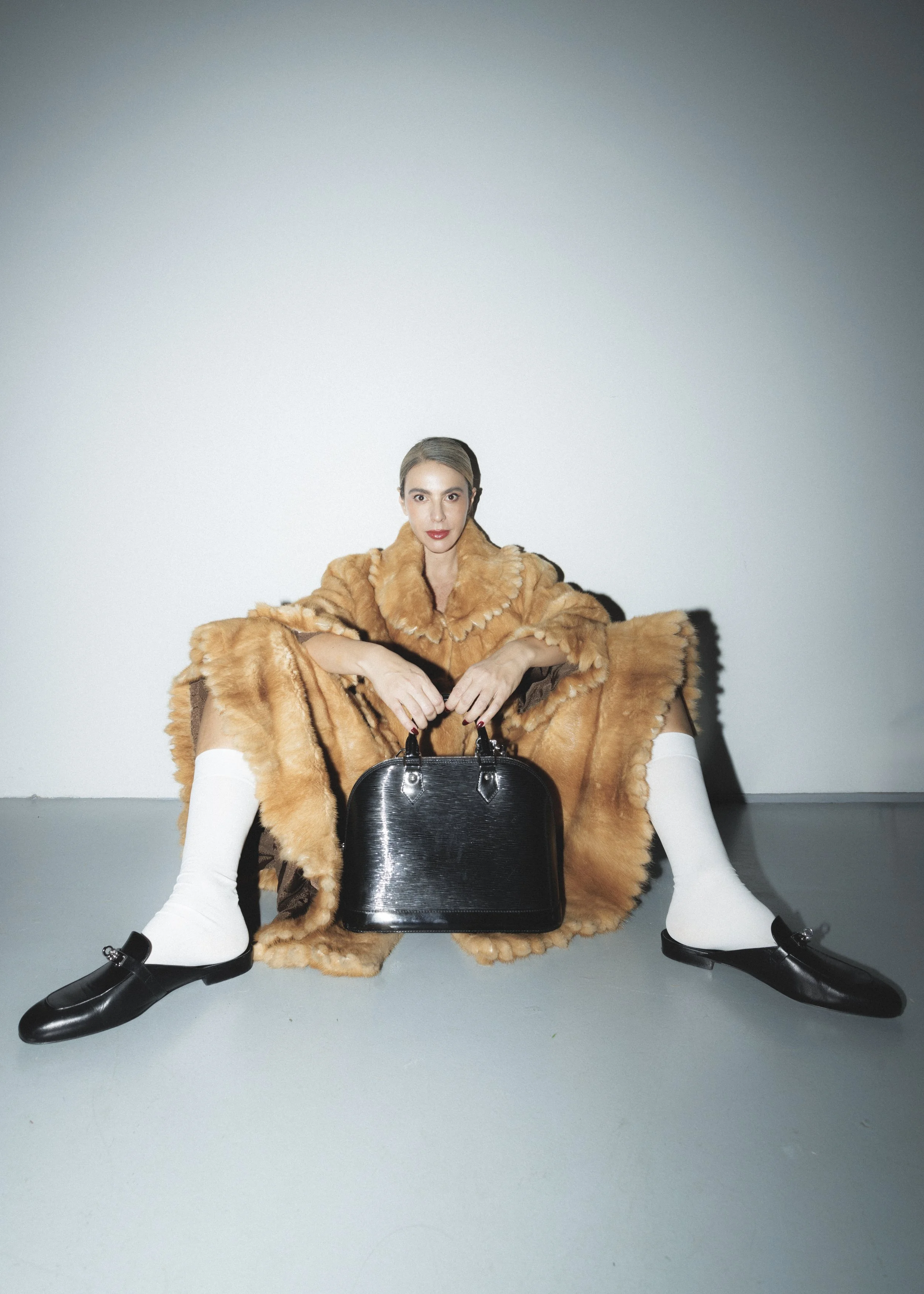 A woman sits on the floor with her legs spread, wearing a long fur coat, white knee-high socks, and black loafers. She holds a black handbag between her legs and looks directly at the camera. The background is plain and light-colored.