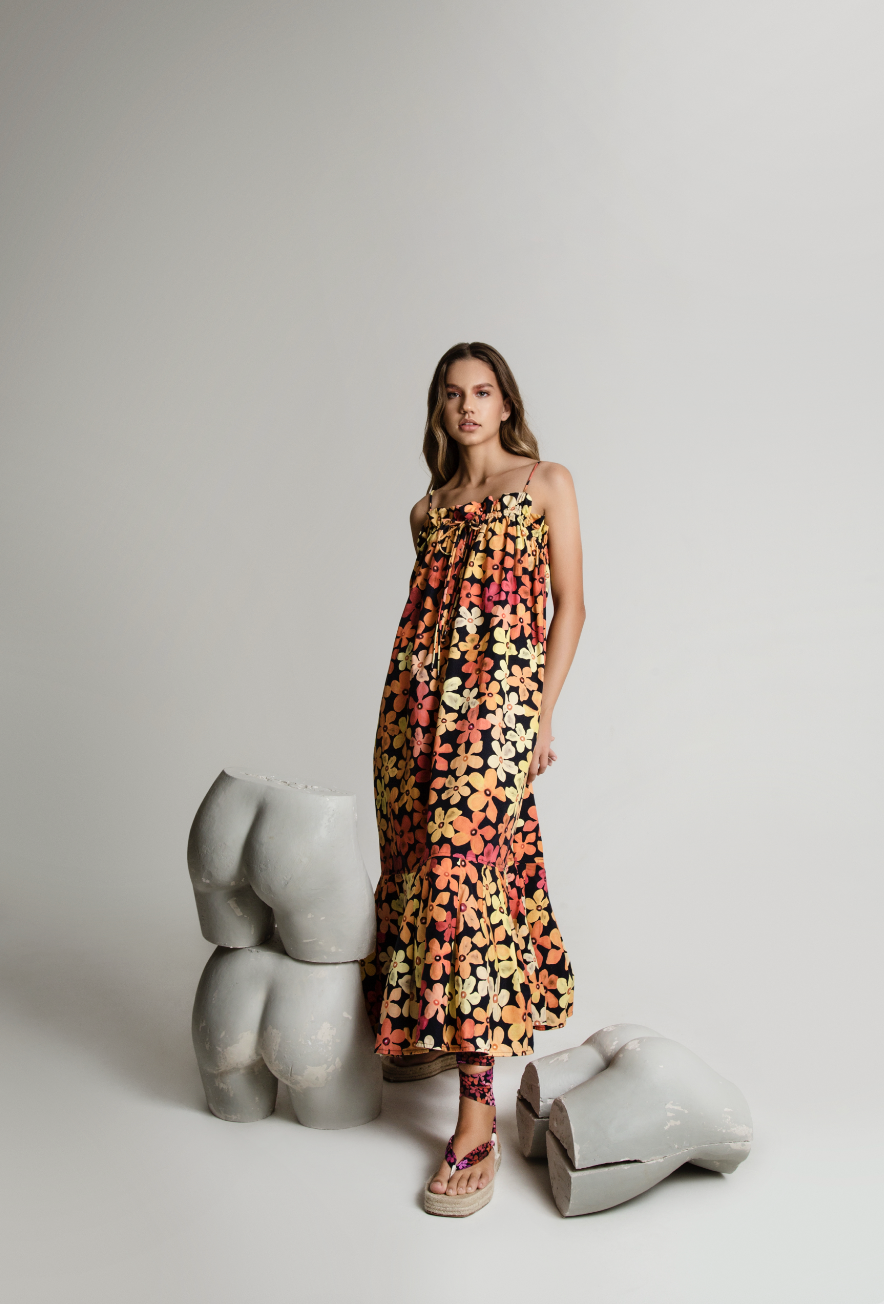 A young woman wearing a colorful floral dress standing in a minimalist studio with abstract shaped sculptures.