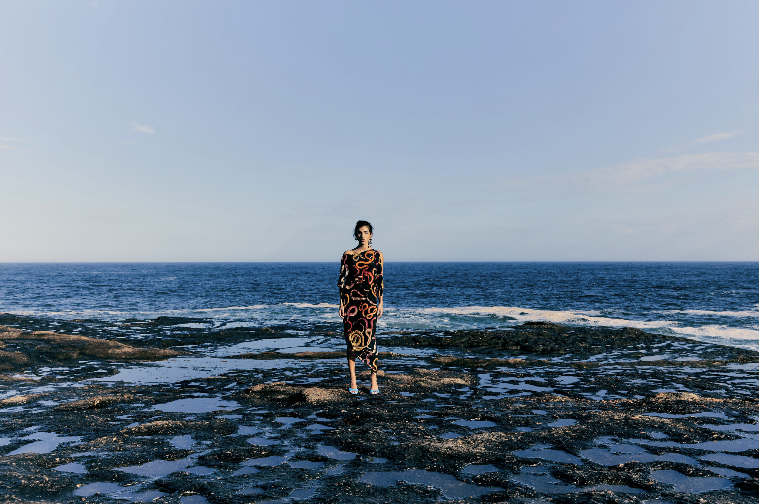 A woman in a long, colorful dress stands on a rocky shore with the ocean and a partly cloudy sky in the background.