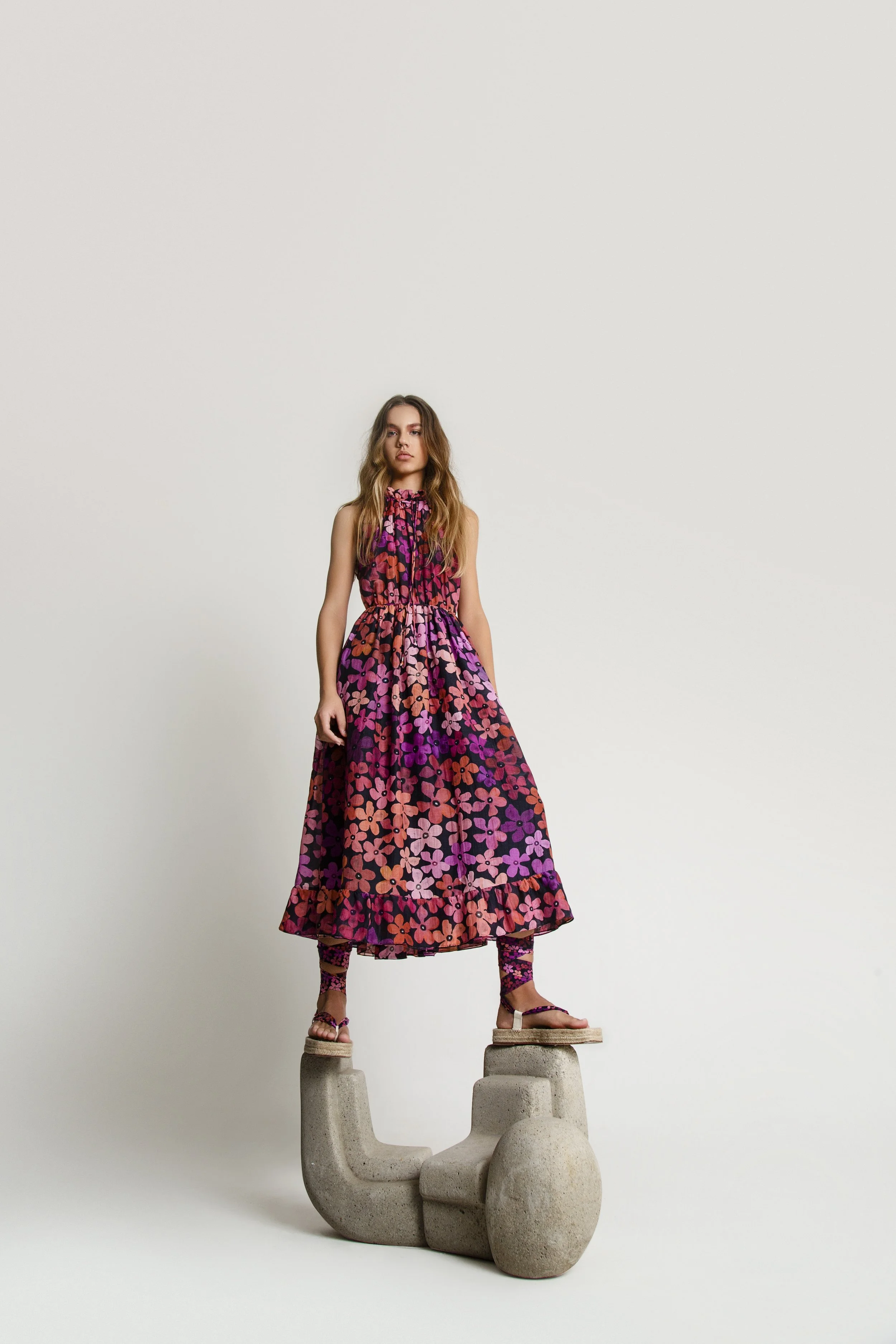 A woman standing on a concrete sculpture, wearing a colorful floral dress and patterned socks, against a plain white background.
