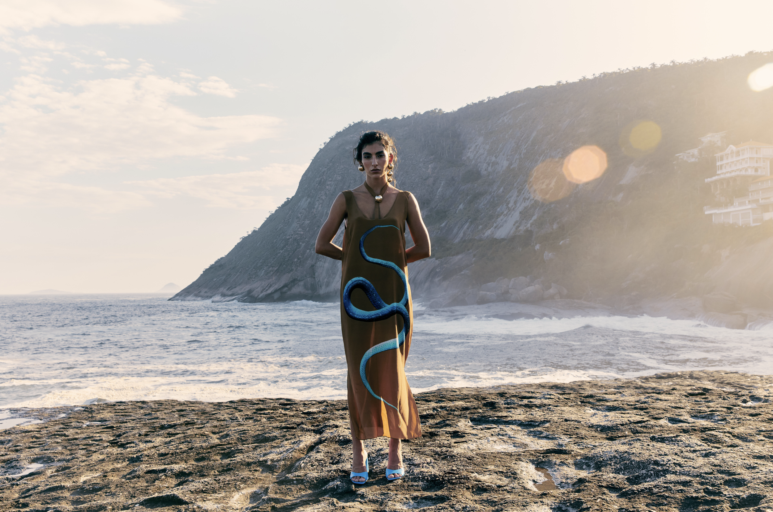 A woman standing on a rocky beach with waves, a large mountain in the background, and buildings on the hillside in the distance, during sunset.