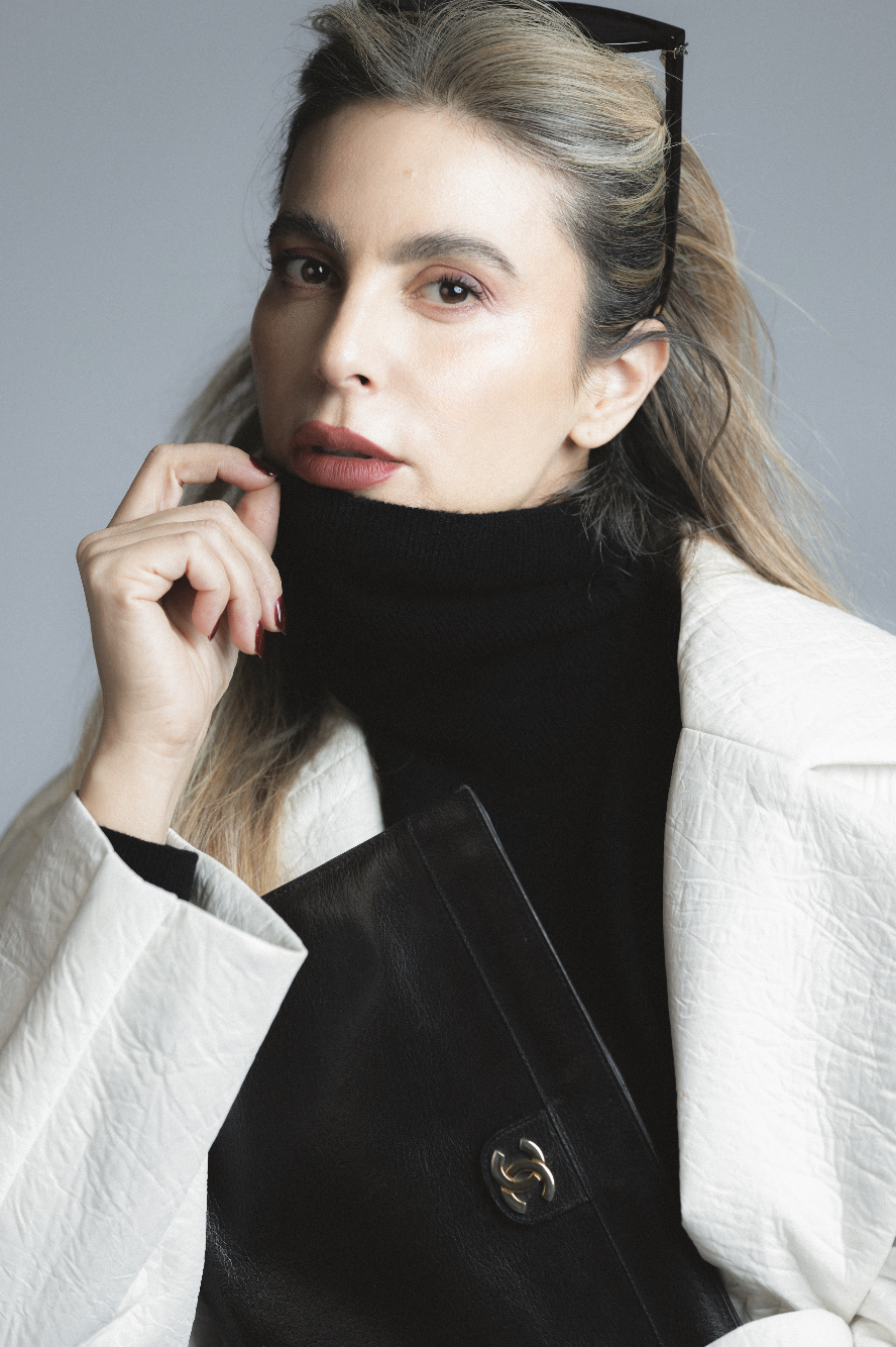 A woman with light skin and long, blonde hair wearing glasses on her head, a black turtleneck, a white textured blazer, and holding a black handbag, looking at the camera against a gray background.