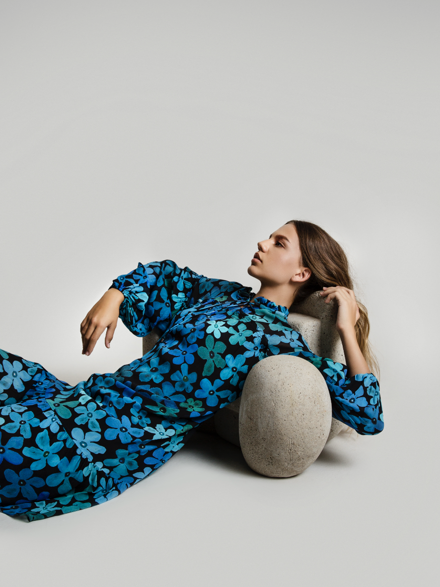 A woman with long wavy hair reclining on a light gray surface, resting her head on a large, oval-shaped stone, wearing a blue floral patterned dress.