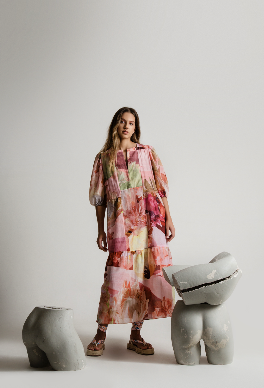 A young woman wearing a colorful, floral-patterned dress standing amidst modern, abstract sculptural pieces against a plain, off-white background.