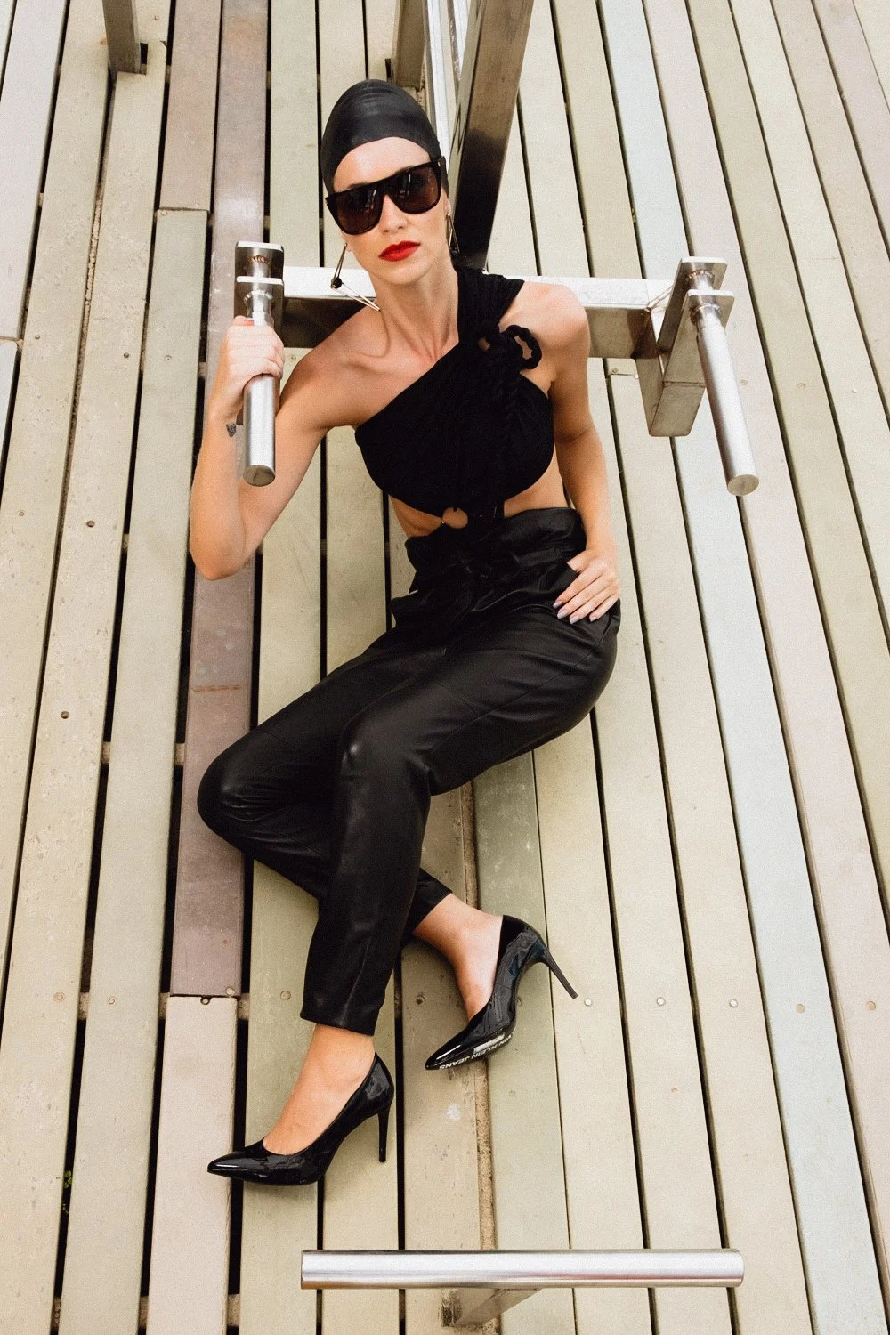 Woman dressed in black, wearing sunglasses and high heels, lying on a wooden deck, holding onto an outdoor exercise machine.