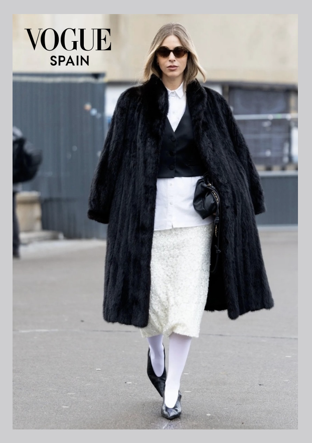 A woman walking outdoors wearing a black fur coat, white skirt, white tights, black shoes, sunglasses, and carrying a small black purse, with a "VOGUE SPAIN" logo in the top left corner.