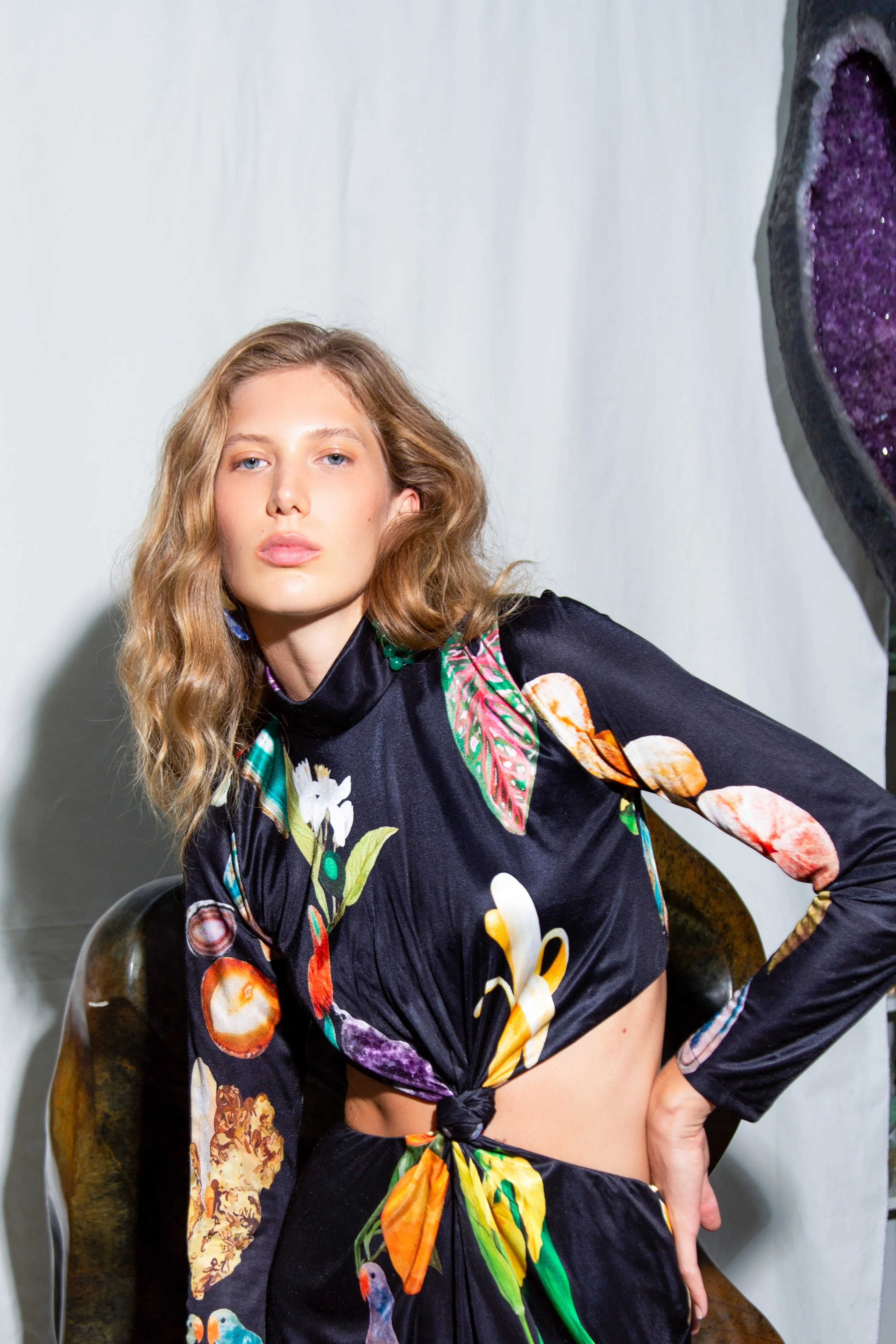 A woman with wavy blonde hair and blue eyes wearing a black floral kimono-style dress with a tie at the waist, sitting on a clear chair against a plain white background.