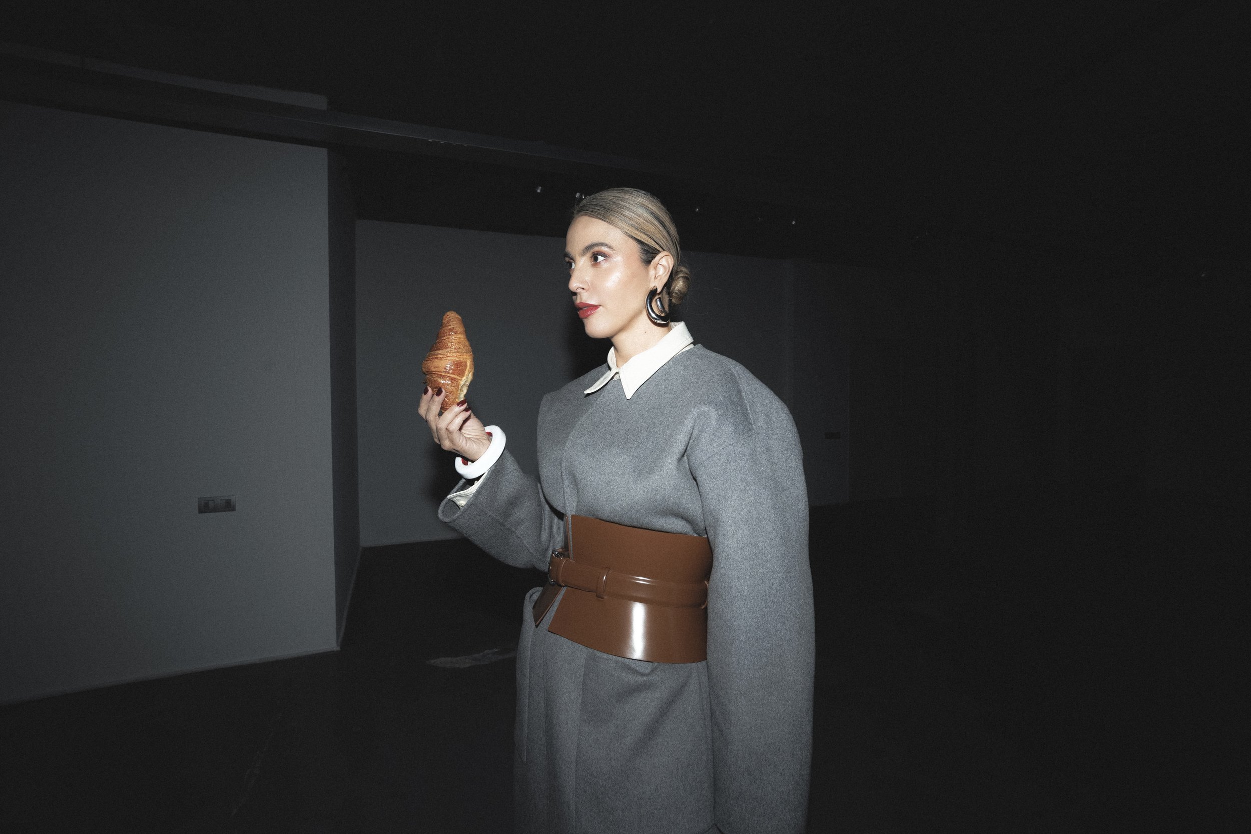 A woman with slicked-back hair, wearing a gray suit with a white shirt and a wide brown belt, holding a croissant in her left hand, standing in a dark room with plain gray walls.