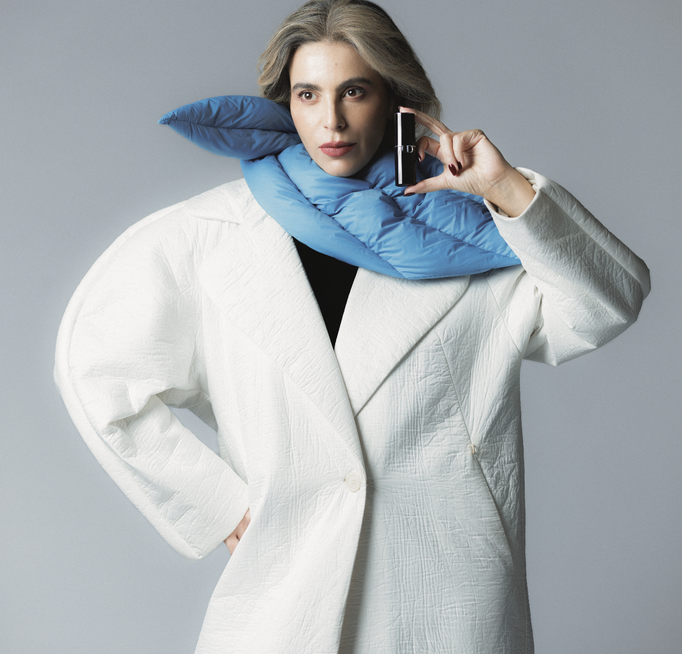 A woman with gray hair breaking a lipstick. She is wearing a white textured blazer and a blue padded scarf around her neck, set against a plain gray background.