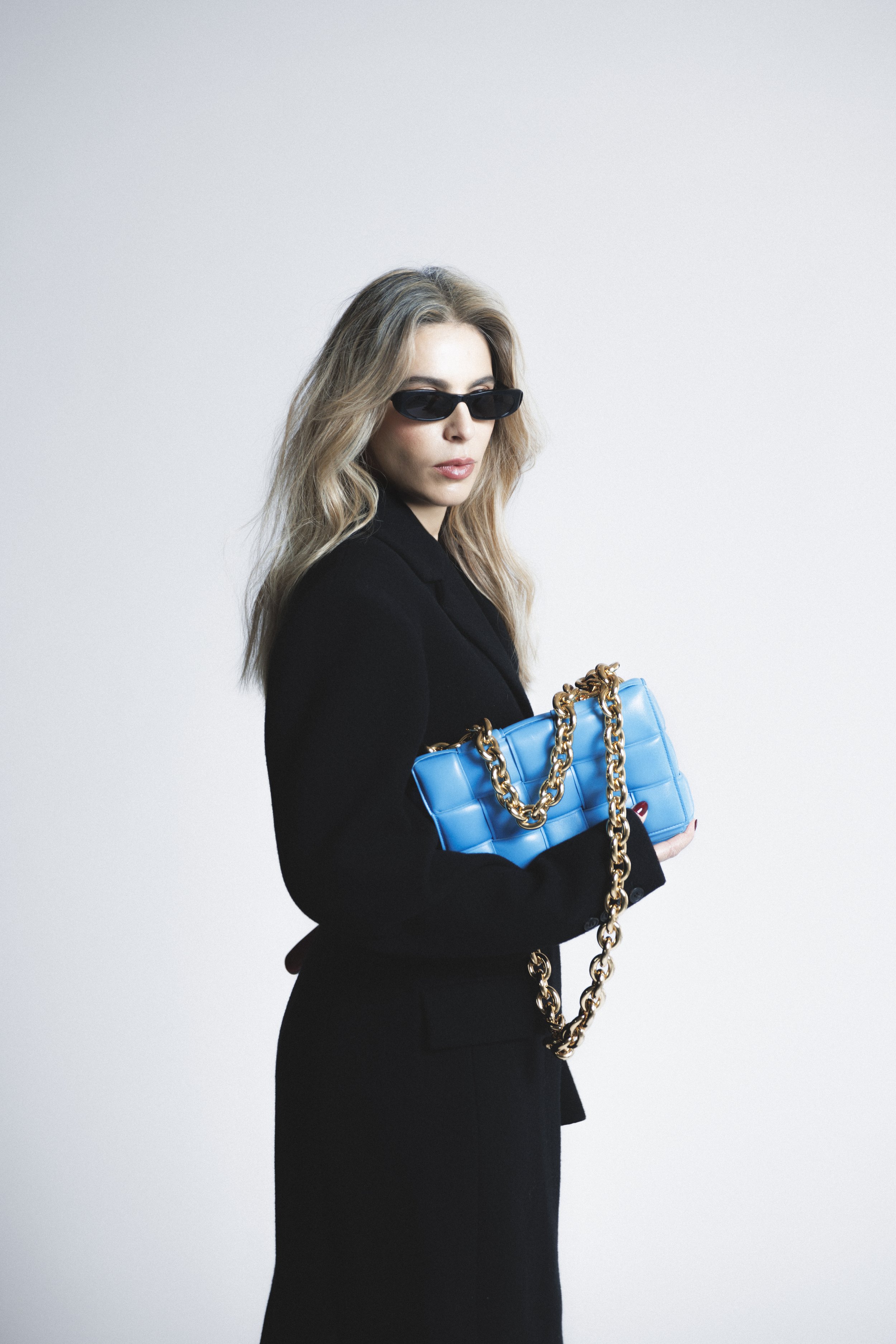 A woman with long, wavy blonde hair wearing black sunglasses and a black outfit is holding a bright blue handbag with large gold chains, standing against a plain white background.