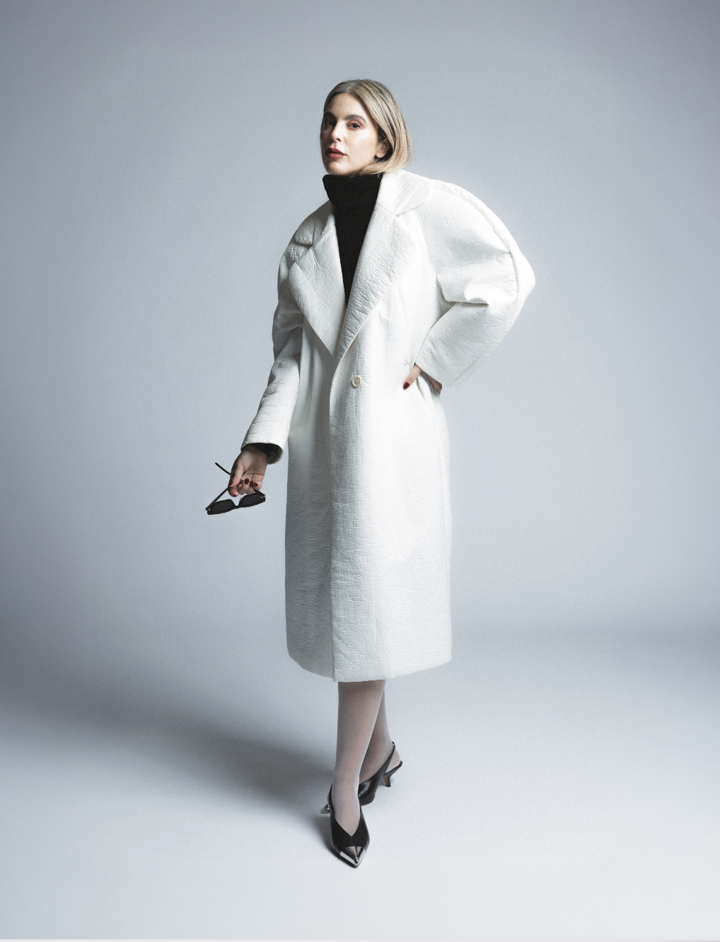 Woman standing against a gray background, wearing a white long coat over a black turtleneck, holding sunglasses in her right hand, and wearing black pointed heels.